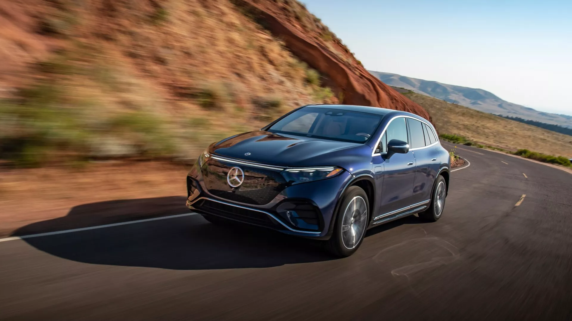 Dynamický snímek modelu Mercedes EQS-SUV 2022, zachycený ze tříčtvrtečního úhlu zleva vpředu, který ukazuje vozidlo v pohybu. Mezi hlavní rysy patří elegantní levá strana vozu, přední kapota, světlomety s pokročilou LED technologií, mřížka, přední nárazník a litá kola. Aerodynamický design karoserie naznačuje luxus a pokročilé inženýrství, což zdůrazňuje výkon a jízdní zážitek. Obrázek vyzařuje pocit svobody a výkonu na čistých silnicích obklopených přírodou.