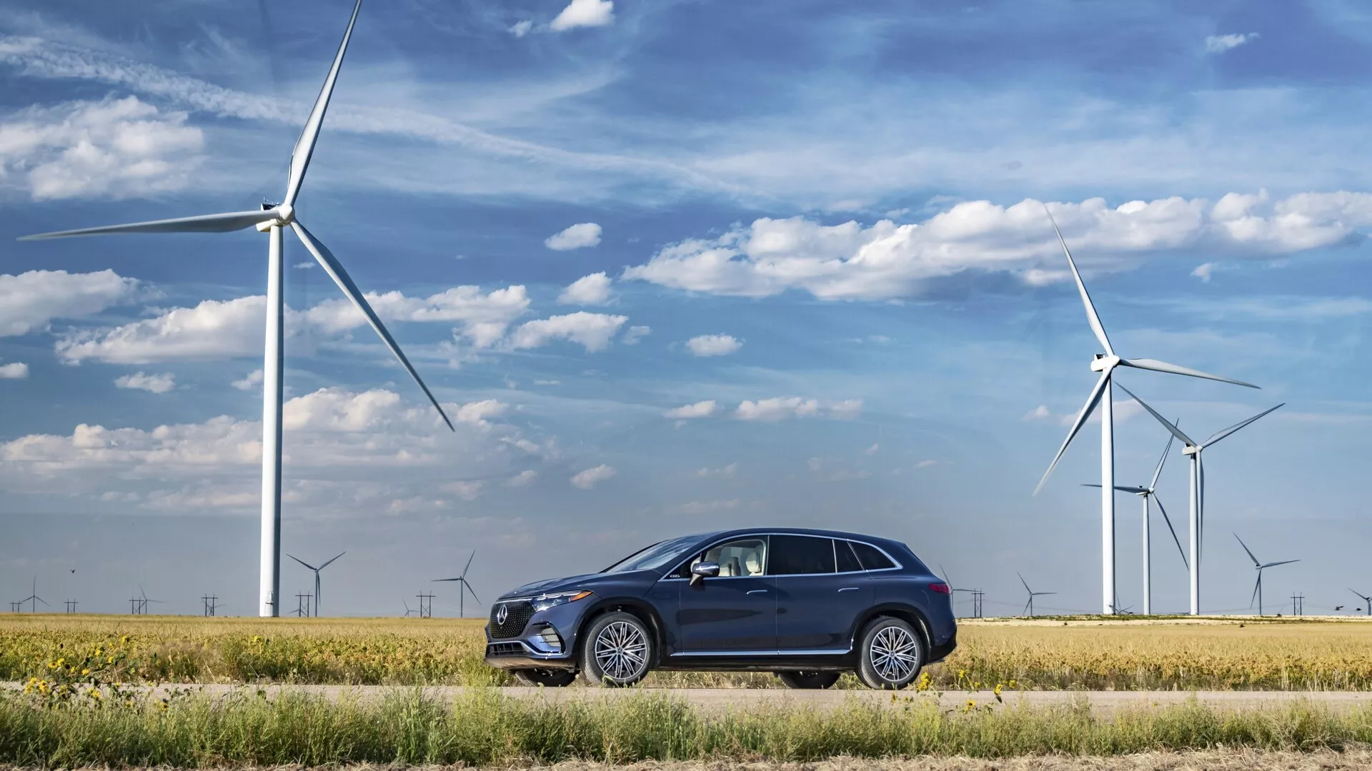 Mercedes EQS-SUV 2022 zaparkovaný v ekologické krajině s větrnými turbínami v pozadí, předvádějící moderní, elegantní design s tmavým zbarvením, vícepaprskovými koly, zapuštěnými klikami dveří, chromovanými rámečky oken, hranatými LED světlomety a integrovanými směrovými světly ve zpětných zrcátkách.