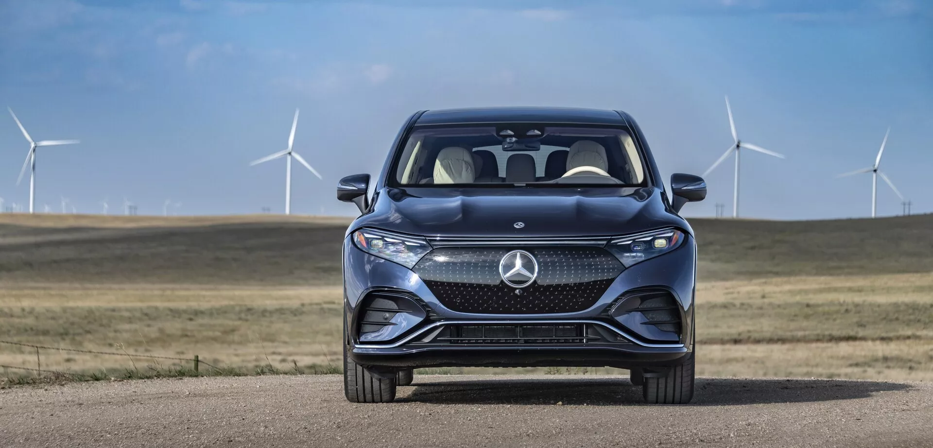 Přední pohled na Mercedes EQS-SUV 2022 proti pozadí jasné oblohy a větrných turbín. Vůz má prominentní masku chladiče s velkým stylizovaným logem a složitým vzorem, který zřejmě obsahuje senzory pro systémy asistence řidiči. Viditelné jsou štíhlé horizontální LED světlomety se zabudovanými denními světly. Sportovní přední nárazník zahrnuje velké přívody vzduchu a kontury kapoty se svažují dopředu pro lepší aerodynamiku. Částečný pohled na přední čtvrtinu odhaluje část blatníků, typickou pro prémiový design. Reflexní čelní sklo a částečně viditelné zrcátko na straně spolujezdce naznačují pokročilé funkce jako směrová světla a monitorování mrtvého úhlu. Pokročilé systémy asistence řidiči a náznaky ekologické jízdnosti jsou naznačeny prostředím.