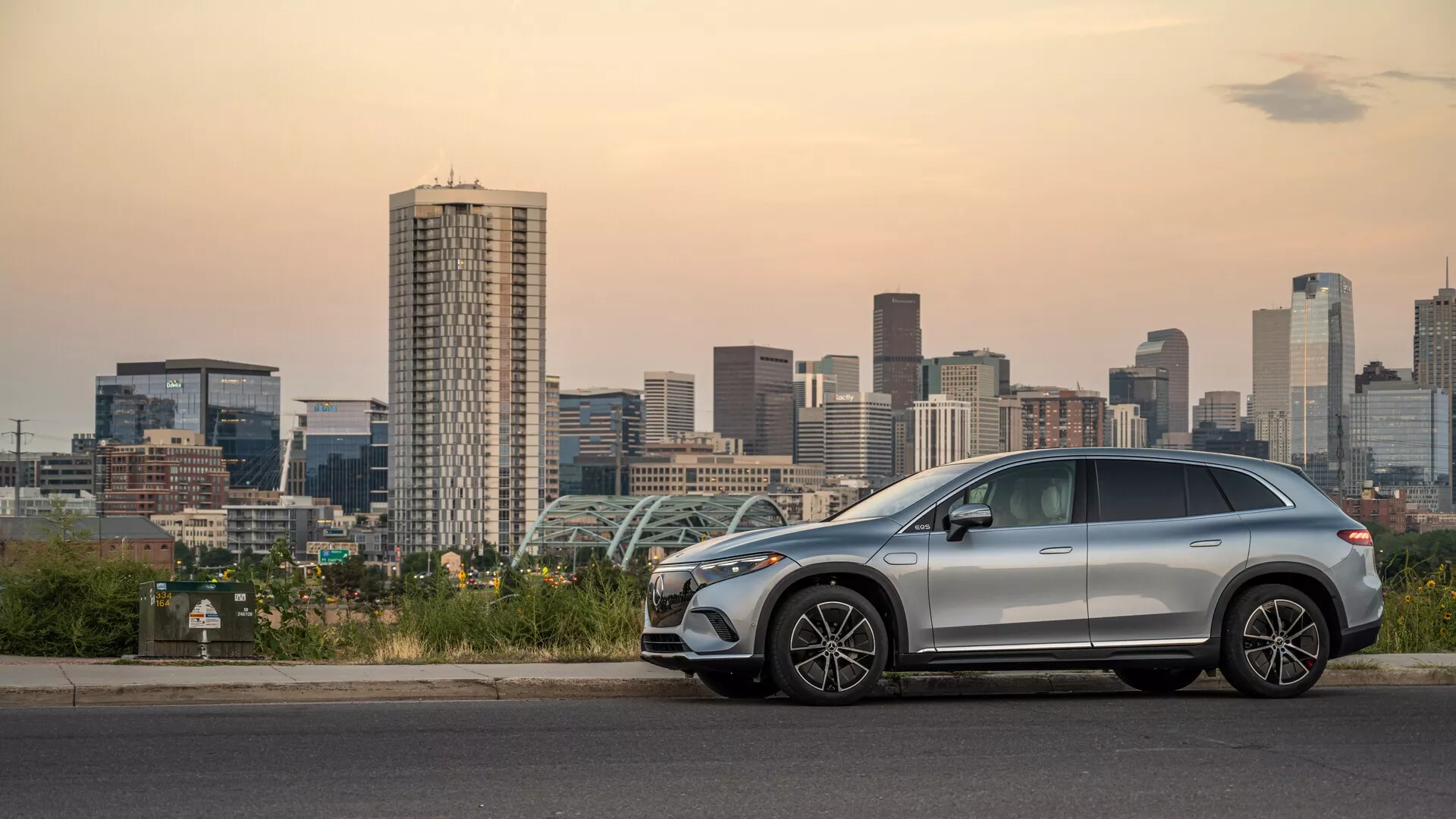 Boční pohled na moderní středně velký Mercedes EQS SUV 2022 zaparkovaný u městského panoramatu za soumraku, předvádějící elegantní aerodynamický design, hliníková kola, střešní nosiče, možnou střešní okno a pokročilé funkce jako integrované směrovky a bezklíčový vstup.