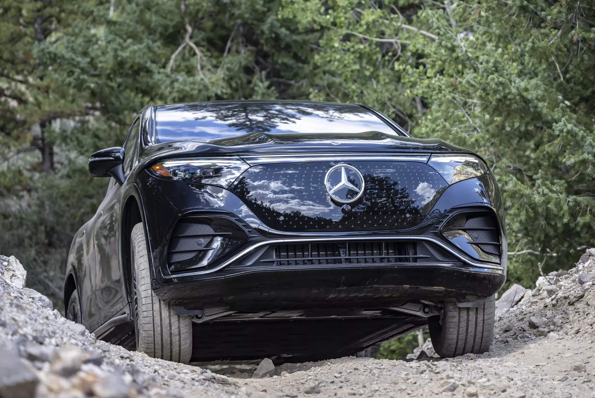 Záběr z nízkého úhlu na černý Mercedes-Benz EQS SUV 2022, zdůrazňující jeho tvarovanou kapotu, velký centrální znak hvězdy, propracovanou mřížku chladiče, elegantní LED světlomety a spodní přívody vzduchu. Vozidlo je zachyceno v přírodním venkovním prostředí, umístěné na svahu, což zdůrazňuje jeho vyšší světlou výšku a potenciál pro jízdu v terénu. Vozidlo je čisté s několika skvrnami od špíny, což podtrhuje jeho lehké terénní využití a pokročilý automobilový design.