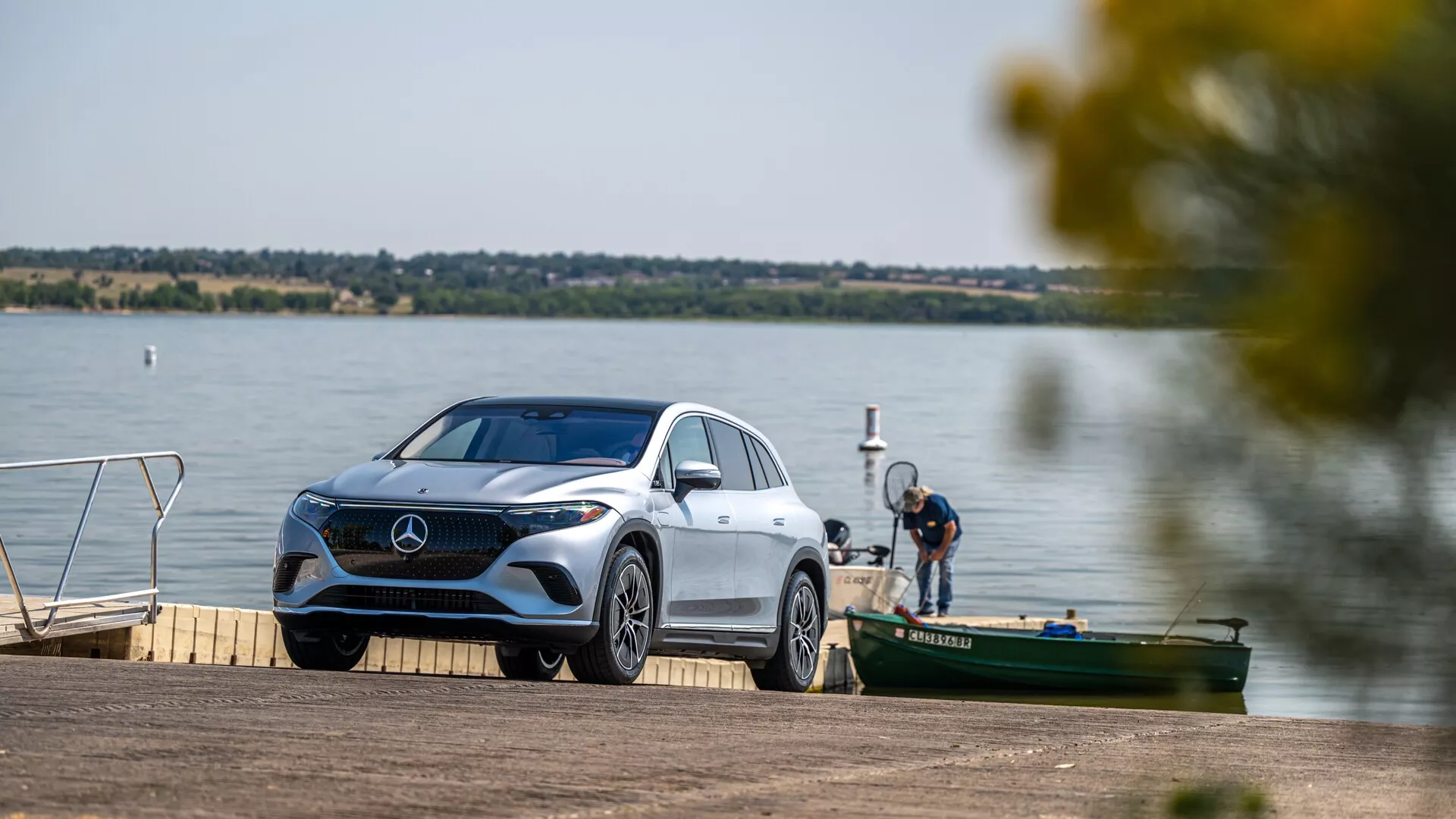 Obrázek zobrazuje luxusní Mercedes-Benz EQS SUV 2022 zaparkovaný u jezera nebo na říčním molu. Vozidlo je mírně nakloněné, což umožňuje pohled z tří čtvrtin přední části, který zvýrazňuje charakteristickou masku chladiče, detailní LED světla, přední nárazník, čelní sklo a část zadní sekce. Prostředí je klidné s vodní plochou a dalšími plavidly v pozadí, což naznačuje rekreační kontext. Scéna je dobře osvětlená, což zvýrazňuje elegantní designové linie a tvary vozu, s rozmazanými listy vytvářejícími přirozený rám.