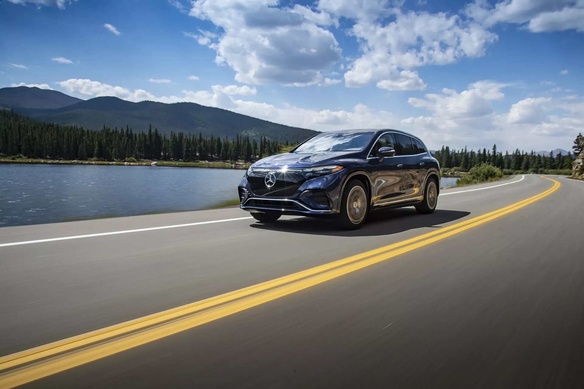 Mercedes EQS SUV 2022 jede po asfaltové silnici s dvojitými žlutými čárami pod částečně zataženou oblohou. Vozidlo je zachyceno z nízkého předotřetinového pohledu, vyniká prominentní přední mřížkou s velkým emblémem, LED světlomety, elegantními liniemi kapoty, slitinovými koly a bočním zrcátkem s možnými integrovanými směrovými světly. Pozadí obsahuje poklidnou krajinu s vodní plochou, horami a jasným horizontem, zdůrazňující pocit cestování a dobrodružství. Pohyb vozu je zvýrazněn mírným pohybovým rozostřením.