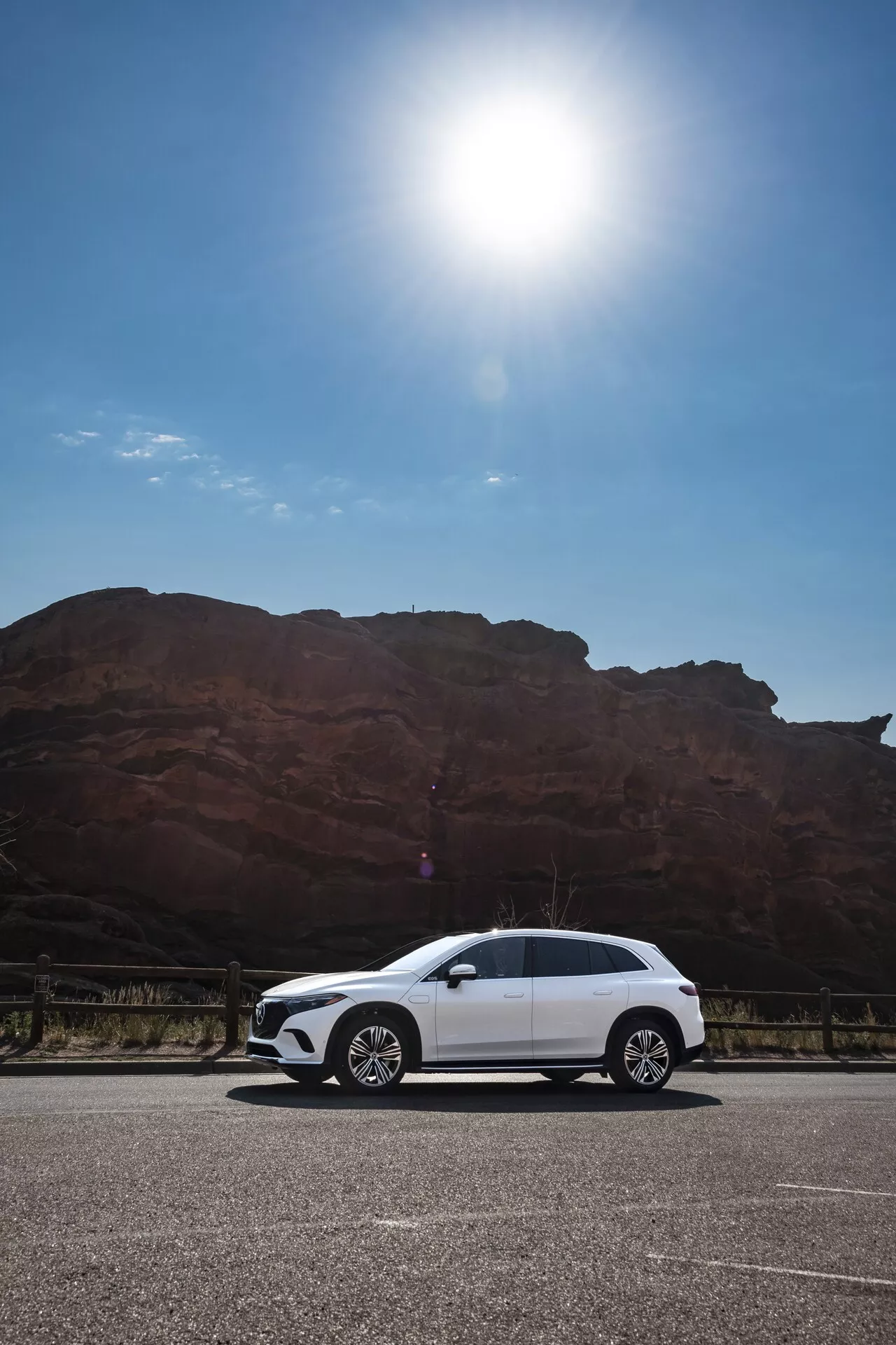 Obraz bílého Mercedesu EQS SUV 2022 zaparkovaného na dlážděné ploše proti velké, červeno-hnědé skalní formaci, pořízený z nízkého úhlu zobrazující boční profil auta včetně klik dveří, bočních zrcátek, ráfků a obrysů karoserie pod jasnou oblohou s odleskem slunce v levém horním rohu.