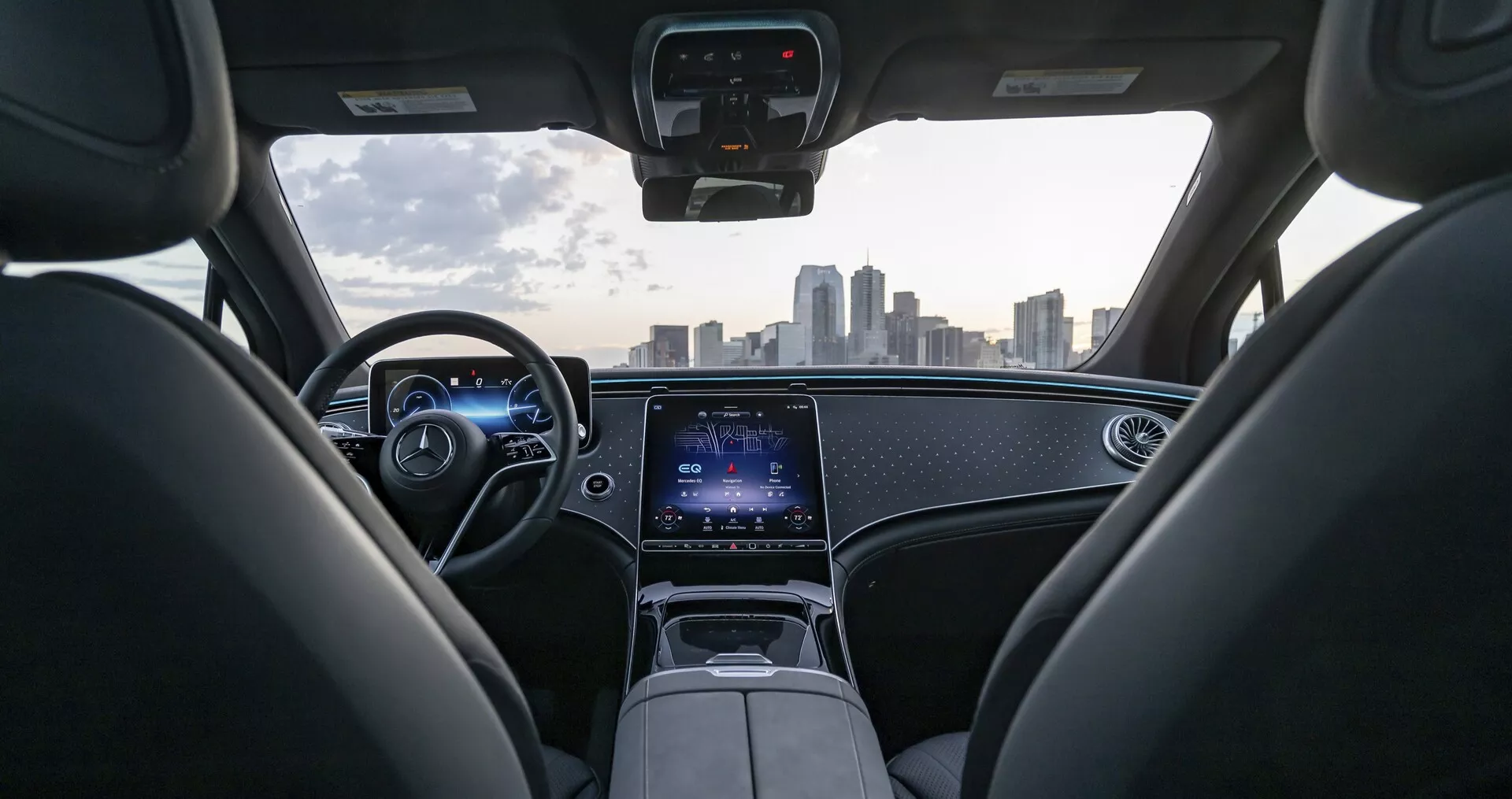 Pohled na interiér vozu Mercedes EQS-SUV 2022 z perspektivy zadního sedadla, ukazující moderní palubní desku s ambientním osvětlením, digitálními přístroji a koženým volantem s integrovanými ovládacími prvky. Středovou konzoli dominuje velká dotyková obrazovka pro infotainment a připojovací funkce. Čelní sklo poskytuje jasný a neomezený výhled na městské panorama, zdůrazňující pokročilou technologii a luxusní design vozu.