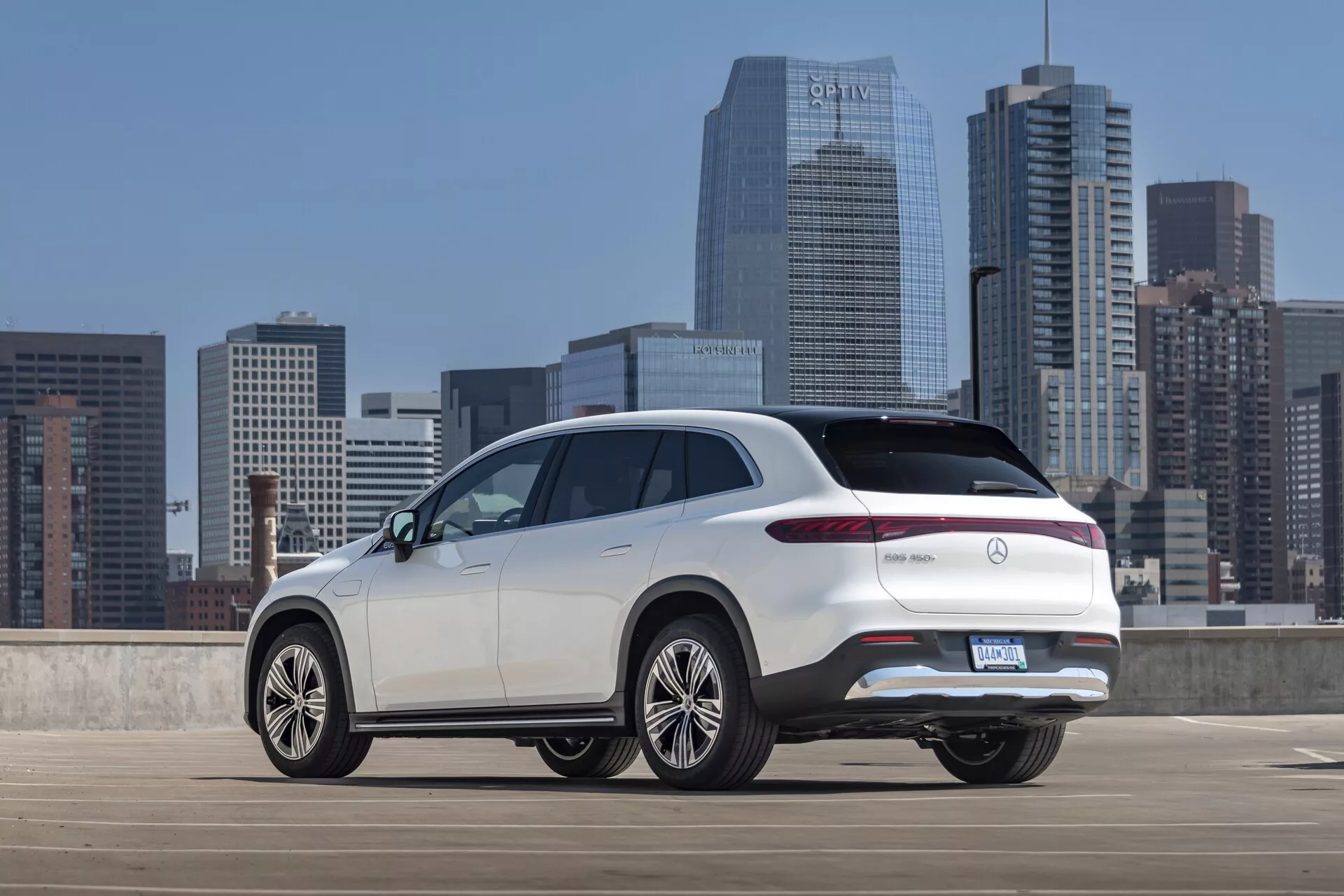 Tři čtvrtě zadní pohled na bílý Mercedes EQS-SUV 2022 z mírně zvýšeného úhlu, který ukazuje horní část zadní části, boční profil a výrazné designové prvky, jako jsou elegantní LED zadní světla, výklopná zadní vrata, zadní nárazník a velká vícepaprsková hliníková kola. SUV je zaparkováno na otevřené městské parkovací ploše s městským panoramatem v pozadí, což zdůrazňuje jeho moderní, luxusní design s potenciálními pokročilými funkcemi typickými pro luxusní modely.