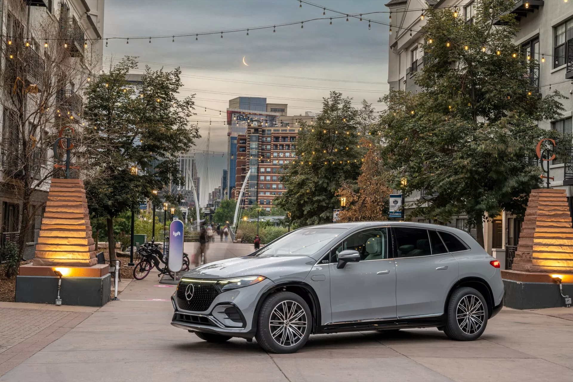 Moderní SUV v městském prostředí během úsvitu nebo soumraku, nízkoúhlý záběr přední čtvrtiny zdůrazňující přední mřížku, světlomety, kapotu a část strany řidiče. Centrální kompozice s květináči a teplým osvětlením ulice přidávají hloubku. Vynikající luxusní prvky zahrnují vícepaprsková slitinová kola, LED světlomety, mřížkový gril s velkým emblémem, integrované zrcátkové směrovky, elegantní designové linie a metalický lak.