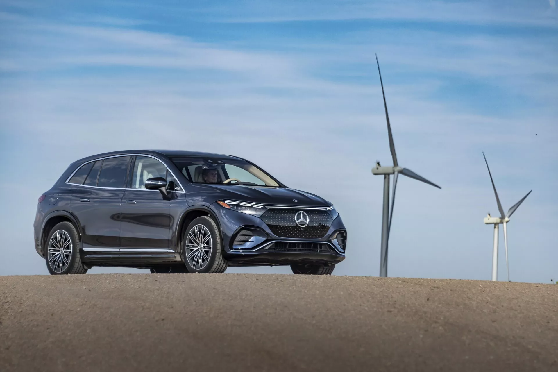 Tříčtvrteční přední pohled na šedý Mercedes EQS-SUV 2022 umístěný na malém kopci, zobrazující moderní design s elegantními liniemi karoserie, výraznými tvary světlometů, výraznou přední maskou s emblémem loga, nehybnými koly a litými koly. Větrné turbíny v pozadí naznačují větrnou farmu nebo venkovskou oblast, což možná poukazuje na ekologické vlastnosti vozidla.