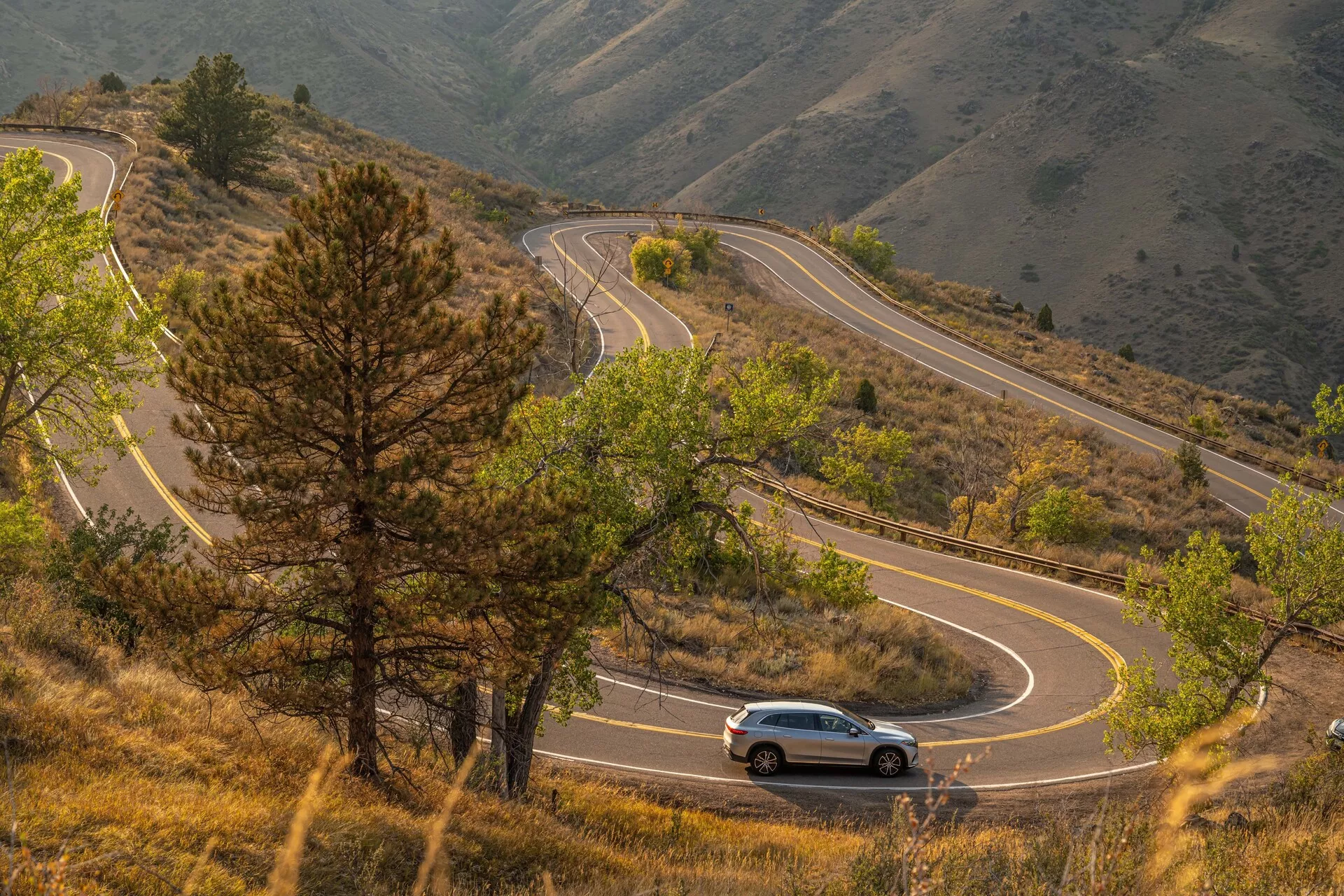 Pohled z ptačí perspektivy na Mercedes EQS SUV 2022 projíždějící klikatou horskou cestou obklopenou kopcovitou, zalesněnou krajinou. Auto je ve středu obrázku, poskytuje boční a mírně zadní pohled, ukazuje designové linie, aerodynamický tvar a viditelné světlomety a zadní světla.