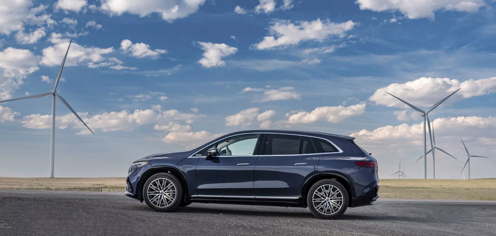 Boční pohled na tmavě zbarvený Mercedes EQS-SUV 2022 zaparkovaný na rovném povrchu silnice, zachycený z pohledu spolujezdce. Elegantní, aerodynamický moderní crossover SUV má pět dveří, velká víceramenná litá kola, panoramatické střešní okno, anténu na střeše a zpětná zrcátka s integrovanými blinkry. Jeho profil naznačuje zvýšený postoj typický pro crossovery. Větrné turbíny v pozadí naznačují ekologická témata.