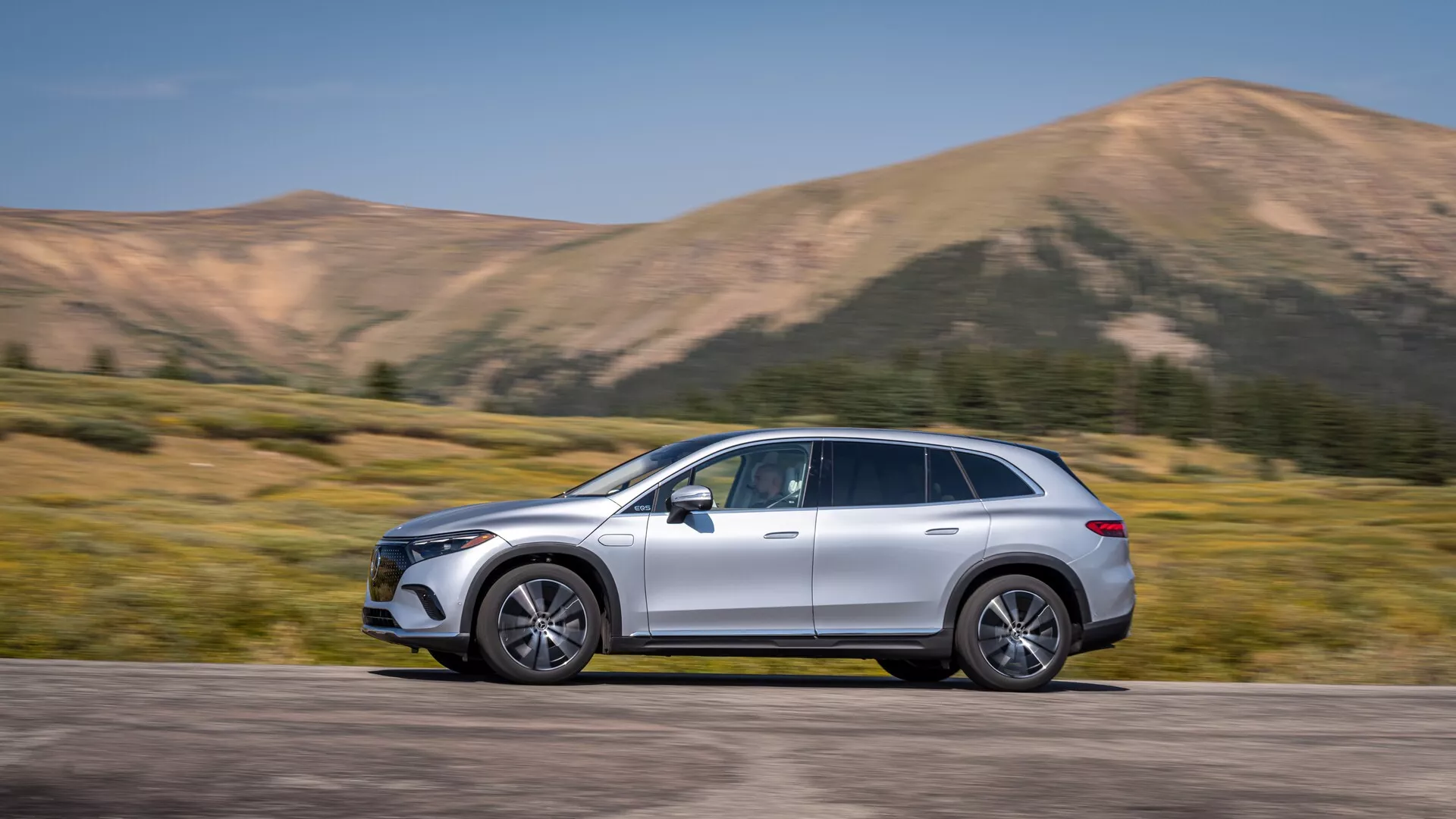 Mercedes EQS-SUV 2022 zachycený v bočním pohledu, umístěný uprostřed rámu s pohybem zřejmým z mírně rozmazaného pozadí. Plnohodnotné SUV s elegantním zakřiveným designem, aerodynamickými bočními zrcátky, litými koly, střešními ližinami, chromovanými lištami a metalickým lakem. Viditelné jsou pokročilé technologické prvky, včetně LED denních světel a zapuštěných klik dveří naznačujících bezklíčový vstup. Horská krajina posiluje dynamický a dobrodružný dojem, ideální pro automobilové inzeráty a životní styl.