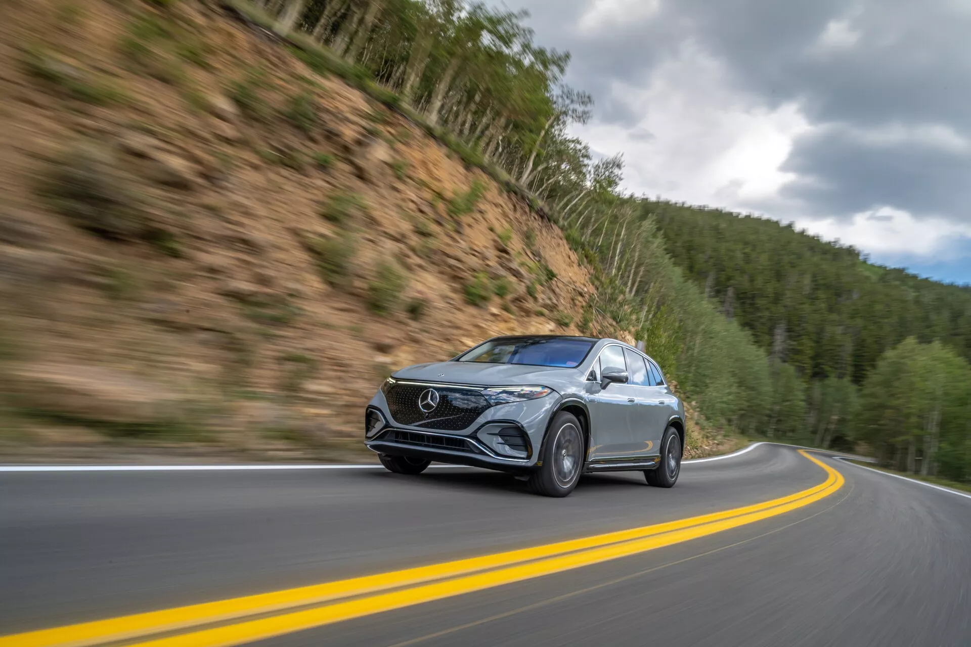 Dynamický snímek vozu Mercedes EQS-SUV 2022 v pohybu na horské silnici, pořízený z nízkého, mírně předního úhlu. Auto, umístěné vlevo od středu, se pohybuje po zakřivené dvouproudové silnici s rozmazaným pozadím, což naznačuje vysokou rychlost. Vybavené elegantními LED světlomety, výraznou přední mřížkou a kovovou barvou, je vozidlo zvýrazněno na pozadí lesem pokrytých hor pod zamračenou oblohou. Detaily jako rozmazaná kola v pohybu a zřetelná boční zrcátka s možnými integrovanými blinkry zvyšují technologický dojem.