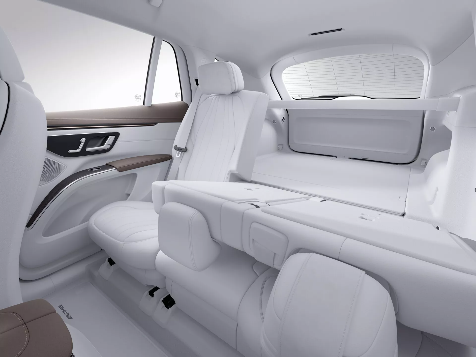 Pohled z výšky na luxusní interiér vozu Mercedes EQS-SUV 2022 s bílým koženým čalouněním, hnědými prvky a dřevěným obložením. Zahrnuje zadní sedadla, část předního sedadla spolujezdce, zadní dveřní panel s ovládáním elektrického okna a malou obrazovku na zadní straně předního sedadla spolujezdce. Jasné osvětlení zvýrazňuje čistý, minimalistický design a dobře udržovaný stav.