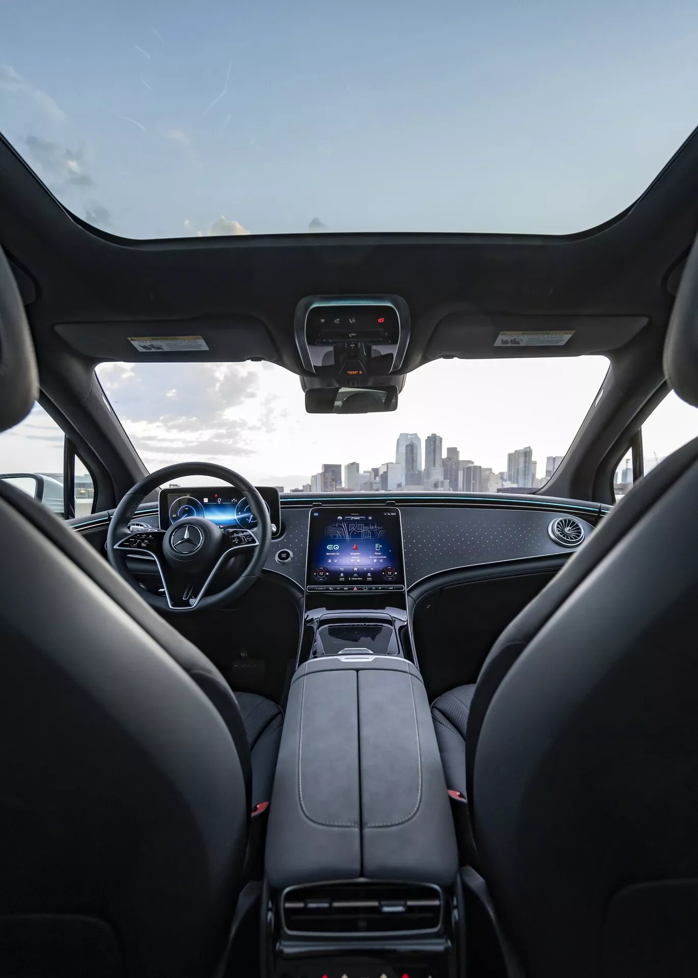 Interiérový pohled na Mercedes EQS SUV 2022 z pohledu z zadního sedadla, ukazující přední sedadla, palubní desku, volant a čelní sklo. Přední sedadla jsou z černé kůže s opěrkami hlavy, palubní deska má elegantní design s kovovými akcenty. Středová konzola obsahuje řadicí páku, držáky nápojů a velký vertikální dotykový displej pro infotainment, GPS a nastavení vozidla. Volant má několik ovládacích tlačítek a za ním je viditelný digitální přístrojový panel. Obrázek také zachycuje městské panorama přes čelní sklo, což naznačuje prémiový, moderní interiér s pokročilou technologií.