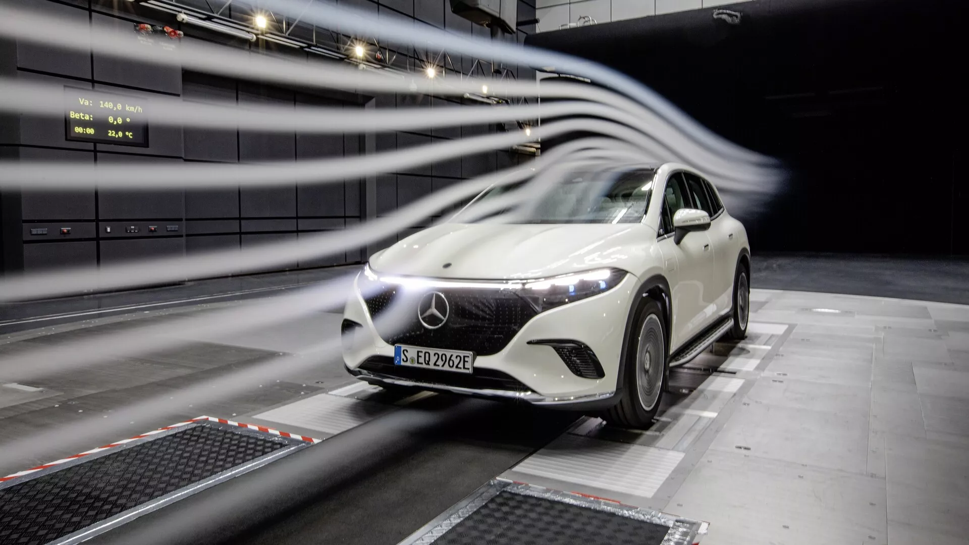 Obrázek bílého vozu Mercedes EQS-SUV 2022 při testování v profesionálním zařízení. Auto, umístěné uprostřed rámce pod mírným úhlem, ukazuje svou přední mřížku, LED světlomety, kapotu, čelní sklo a část strany řidiče. Linie rozmazání pohybu naznačují aerodynamické testování. Futuristický design, velké logo na mřížce a vnitřní testovací prostředí.