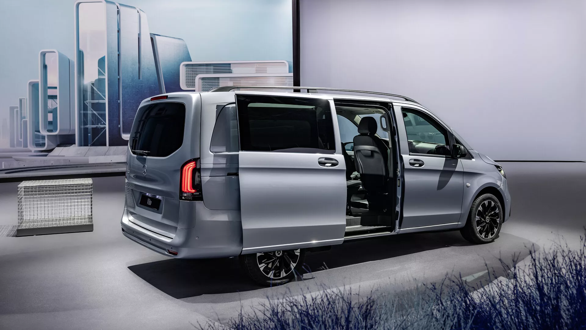 Stříbrný minivan Mercedes-Benz Vito 2024 (447 facelift) se otevřenými posuvnými bočními dveřmi předvádějící elegantní moderní design z tříčtvrtečního zadního úhlu, zdůrazňující aerodynamický profil vozidla, zadní světla LED, slitinová kola a nahlédnutí do pokročilého interiéru s potenciálně prémiovým čalouněním a moderním palubním stolem.
