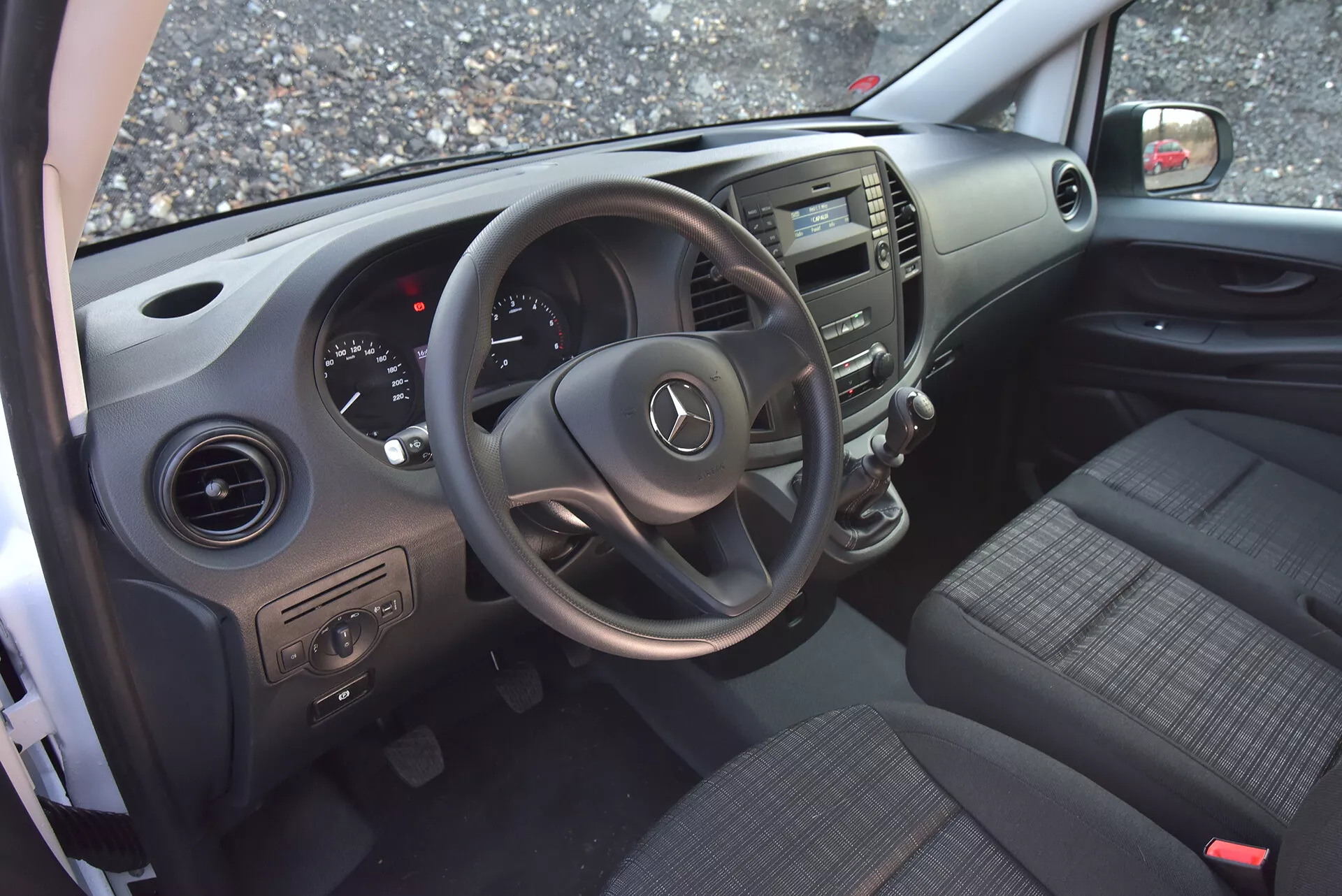Interiér Mercedes-Benz Vito Tourer z roku 2014 viděný ze strany otevřených dveří řidiče, zdůrazňující volant s logem Mercedes, palubní desku, středovou konzolu, sedadla potažená látkou, ovládání na dveřích a větrací otvory.