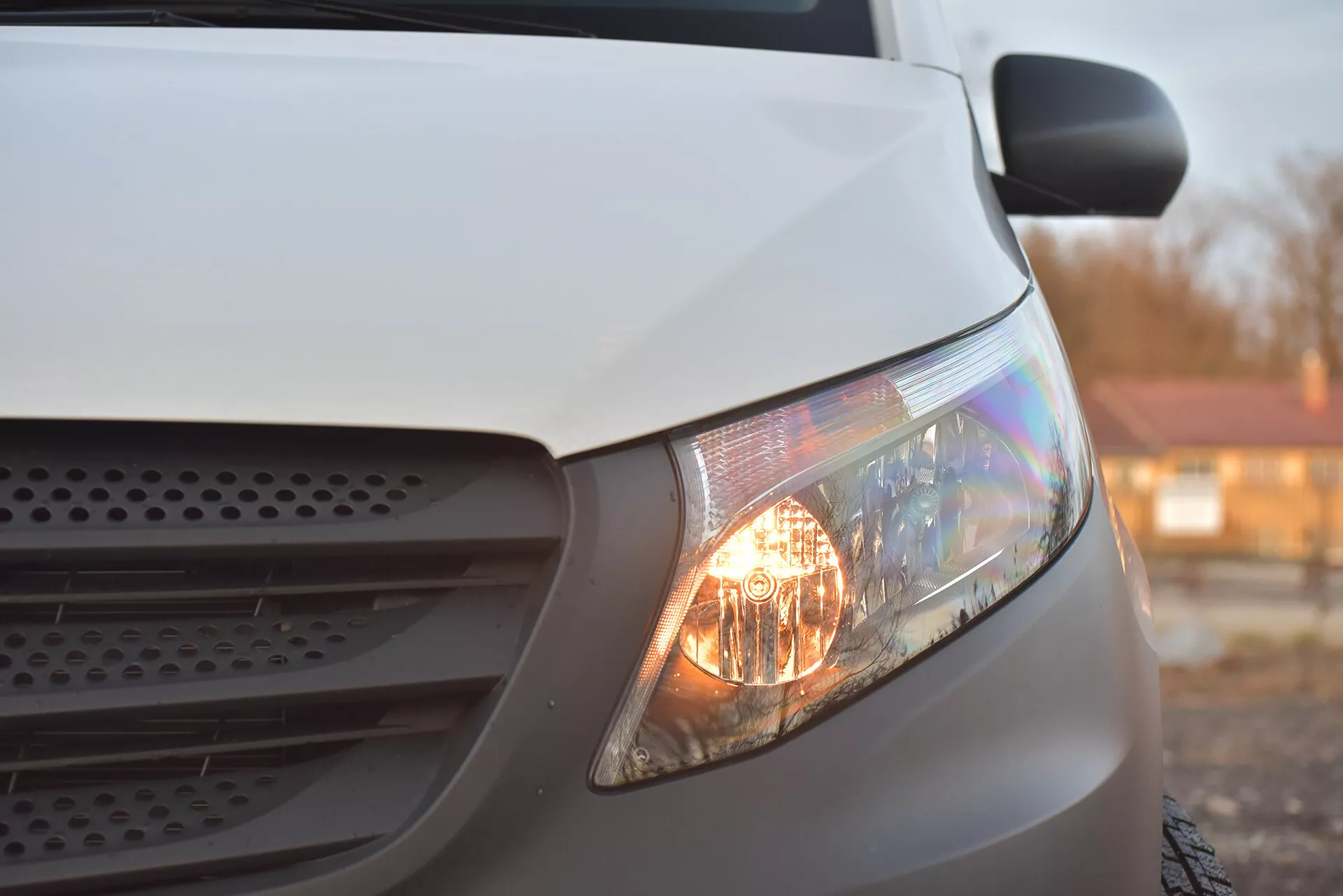 Detailní boční pohled na bílý Mercedes-Benz Vito Tourer z roku 2014 s důrazem na osvětlený světlomet, část mřížky a kapotu, zdůrazňující jeho halogenové světlomety a aerodynamický design ve slabém osvětlení.