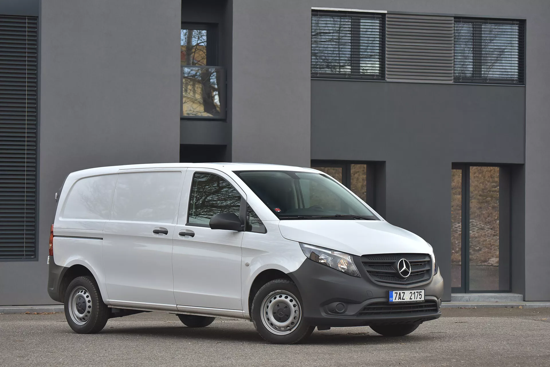 Bílý dodávkový vůz Mercedes-Benz Vito Tourer z roku 2014 zaparkovaný na asfaltové ploše, zobrazení předního a bočního profilu s jasným pohledem na emblém na mřížce a moderní světlomety, fotografováno v zataženém dnešním světle před minimalistickou budovou