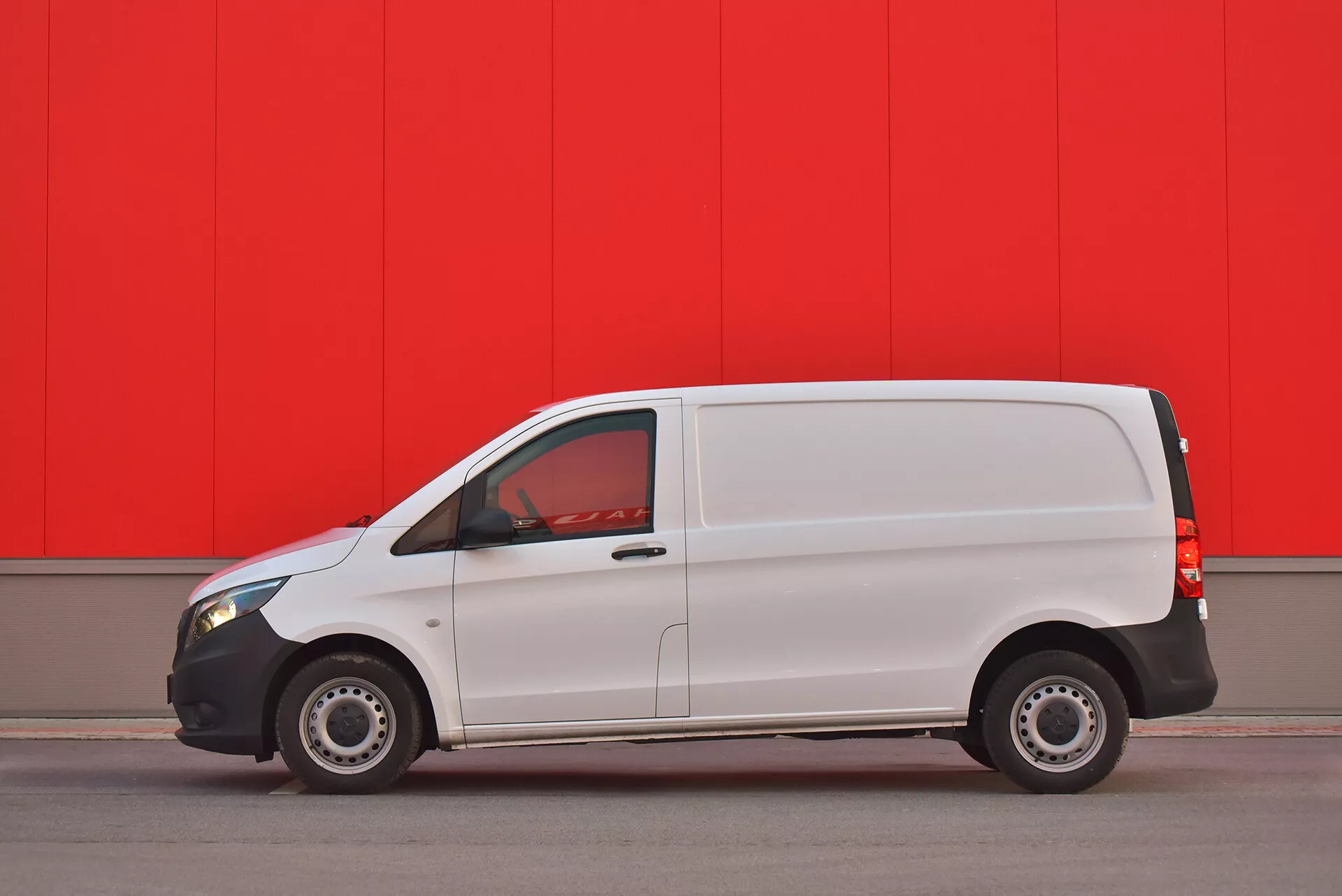 Bílá dodávka Mercedes-Benz Vito Tourer z roku 2014 parkovaná paralelně u živě červené zdi, ukazuje boční profil s viditelnými dveřmi, okny a ocelovými koly, zachycená z nízkého úhlu pro zdůraznění designu a funkcí vozidla