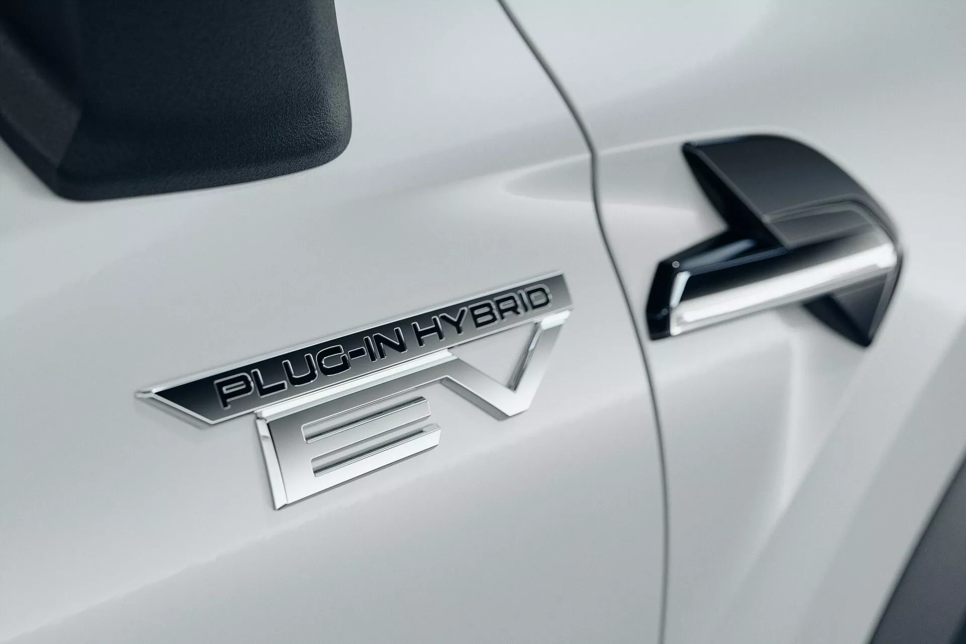 Detailní záběr na bílý automobil Mitsubishi ASX 2023 druhé generace, s výrazným odznakem 'PLUG-IN HYBRID EV', elegantním černým bočním zrcátkem s integrovaným blinkrem a lesklou bílou aerodynamickou karoserií.