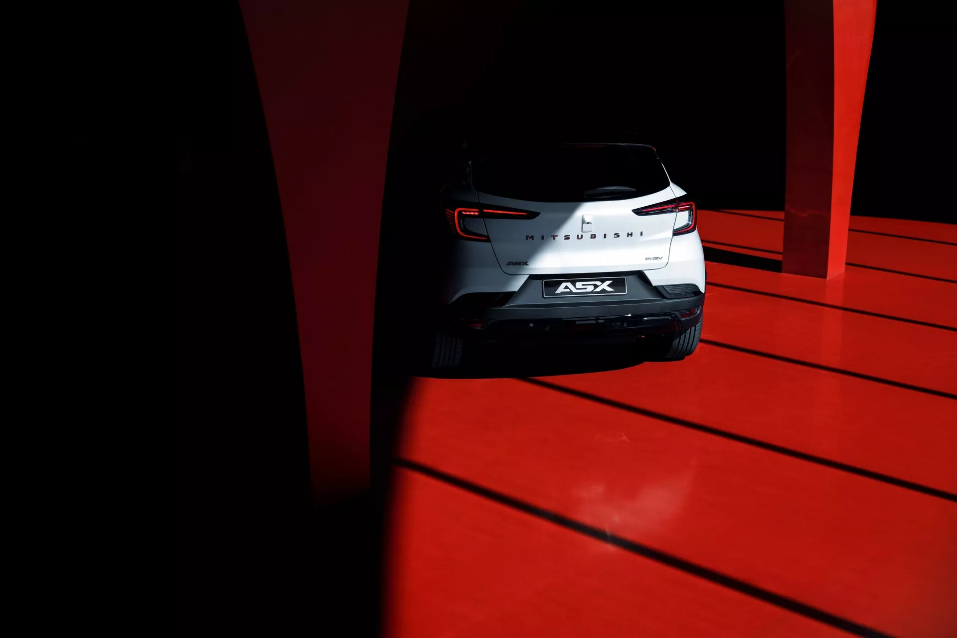Nízký úhel záběru na zadní část kompaktního SUV Mitsubishi ASX 2023 s vypracovanými zadními světly, chromovým znakem a zadním sklem, zachycený za vysokého kontrastu denního světla na živě červeném povrchu.