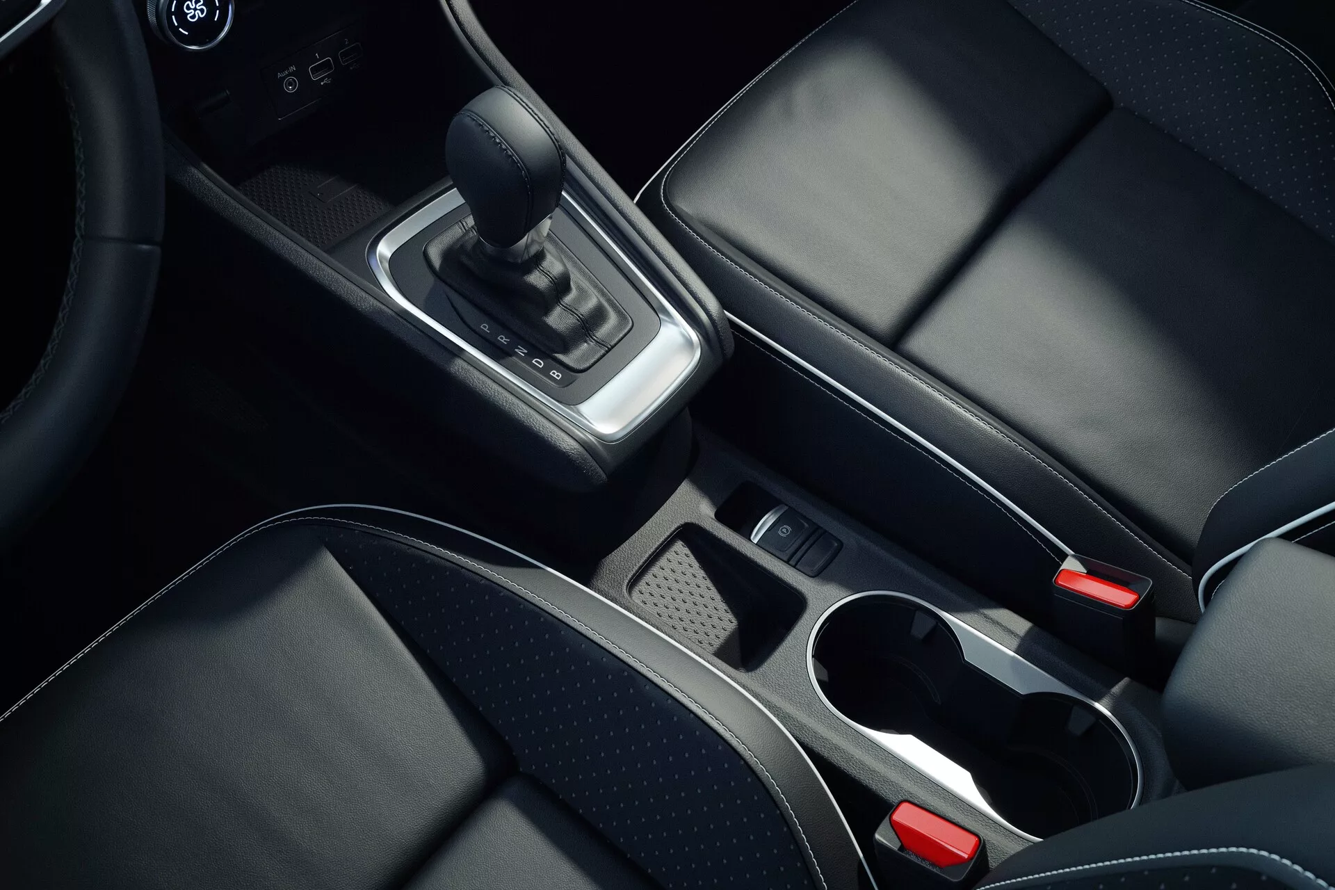 Interiérový snímek středové konzole automobilu Mitsubishi ASX 2023 druhé generace z nadhledu, zaměřený na detaily jako kožená páka řazení, chromové akcenty kolem oblasti řazení, úložný prostor s vyjímatelnou přihrádkou, dva držáky na nápoje s chromovanými lemy, černá kožená sedadla s viditelným šitím, elektronická parkovací brzda a částečně viditelná středová opěrka a systém infotainment nebo klimatizace, představující vysoce kvalitní materiály a moderní design.