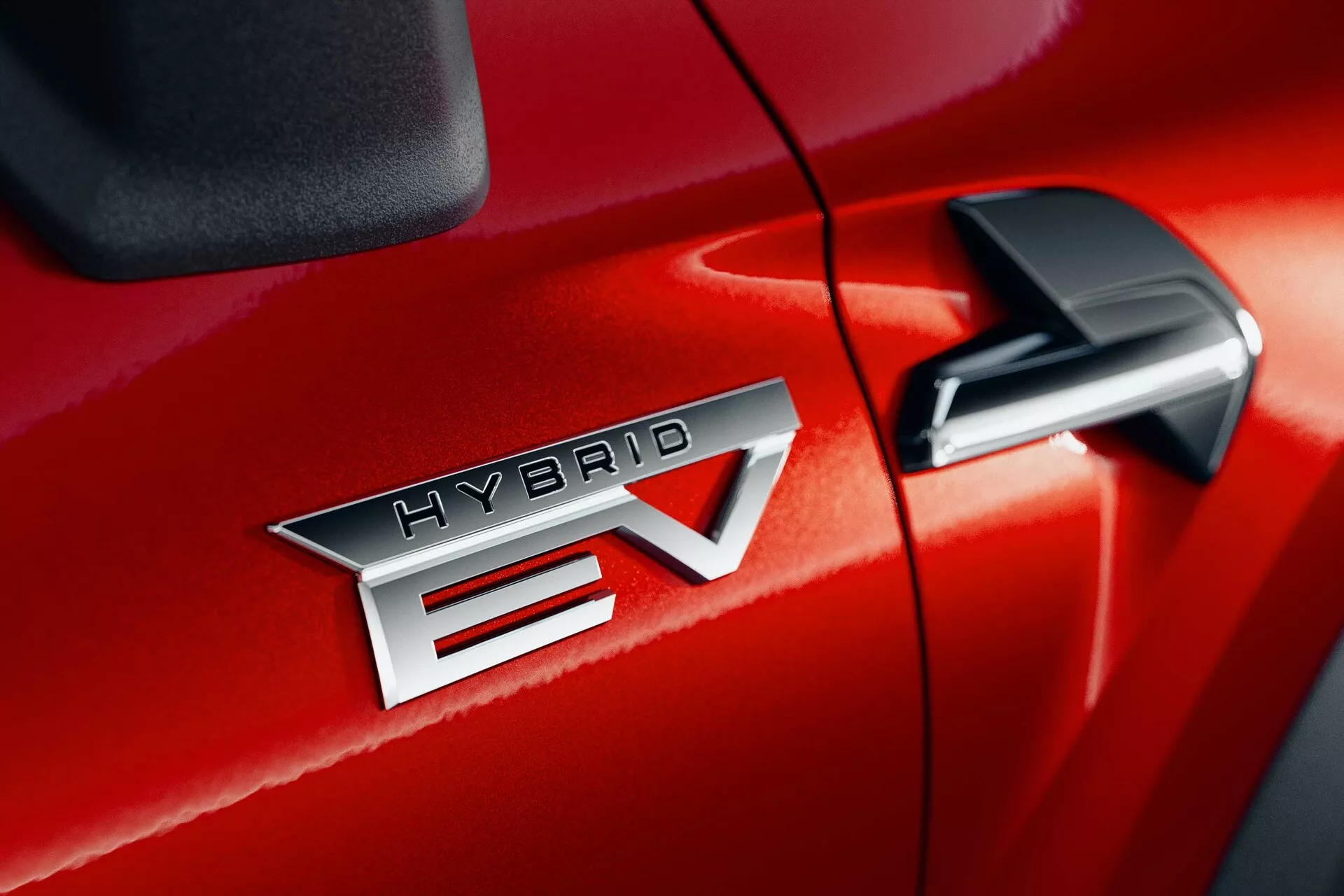 Detailní záběr na Mitsubishi ASX 2023 (2. generace) v červené barvě, ukazující výrazný znak 'HYBRID EV', který naznačuje technologii hybridního elektrického vozidla. Znak 'HYBRID EV' je umístěn ve středu, zdůrazňující kovový finiš a přesné písmo. 'EV' má výrazný design šipky procházející přes 'V'. Boční zpětné zrcátko je částečně viditelné v pravém horním rohu, což naznačuje přítomnost dalších technologických prvků. Lesklá povrchová úprava laku a těsný šev mezi předními dveřmi a čelní částí auta naznačují vynikající kvalitu zpracování.