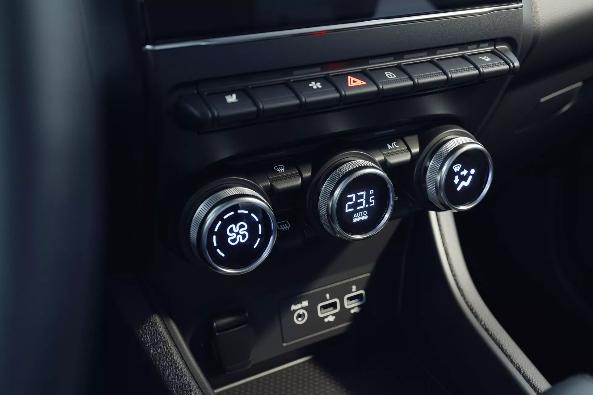 Detailní pohled na interiér klimatizačního systému Mitsubishi ASX 2023 (2. generace). Obrázek pořízený z vyvýšeného úhlu se zaměřuje na středovou konzoli s třemi ovládacími kolečky - levé nastaveno na 'LO' a pravé na '23.5'. Střední kolečko pravděpodobně řídí rychlost nebo směr ventilátoru. Nad nimi jsou tlačítka pro odmlžování, výstražná světla a cirkulaci vzduchu. Pod nimi jsou moderní USB porty. Design je minimalistický s elegantními černými povrchy.