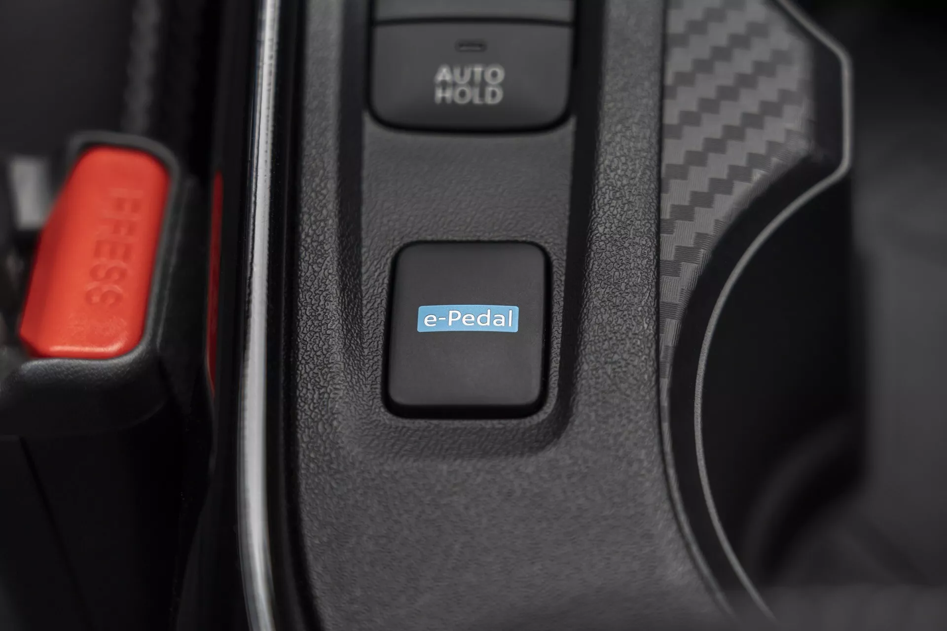 Detail interiéru Nissan Juke (F16) 2019 se zaměřením na systémový spínač e-Pedal pro řízení elektrického nebo hybridního pohonu, s viditelným tlačítkem AUTO HOLD a červenou sponou bezpečnostního pásu naznačující pokročilou automobilovou technologii a prvotřídní interiérové prvky