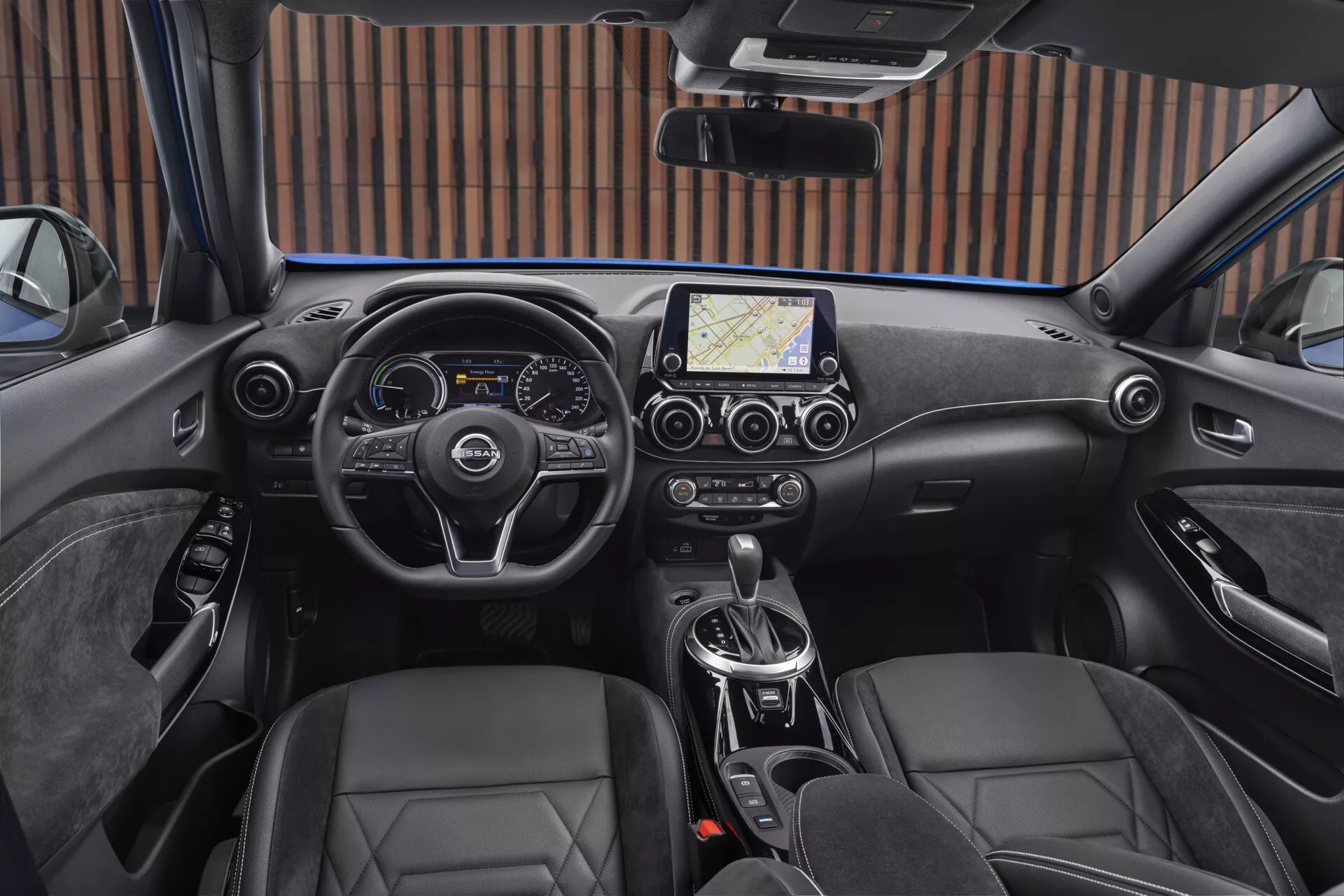 Interiér Nissan Juke F16 z roku 2019 z pohledu řidiče, obsahující moderní volant se třemi rameny s ovládacími tlačítky, kombinovaný digitální a analogový přístrojový panel, centrální dotykový informační systém s navigací, klimatizaci s dvojzónovou regulací, automatickou převodovku, sedadla typu bucket potažená kůží a elektronickou parkovací brzdu.