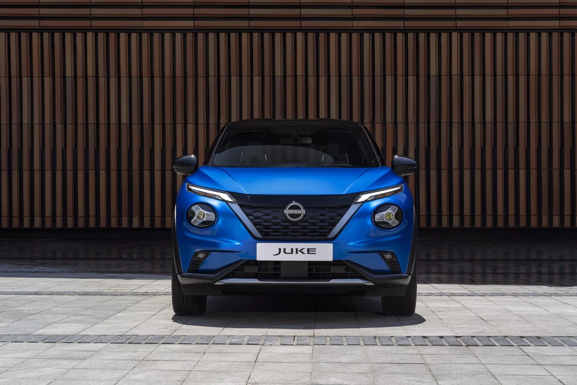Přední pohled na modrý Nissan Juke F16 z roku 2019, předvádějící elegantní LED světlomety, mřížku V-motion a sportovní nárazník na pozadí s horizontálními liniemi