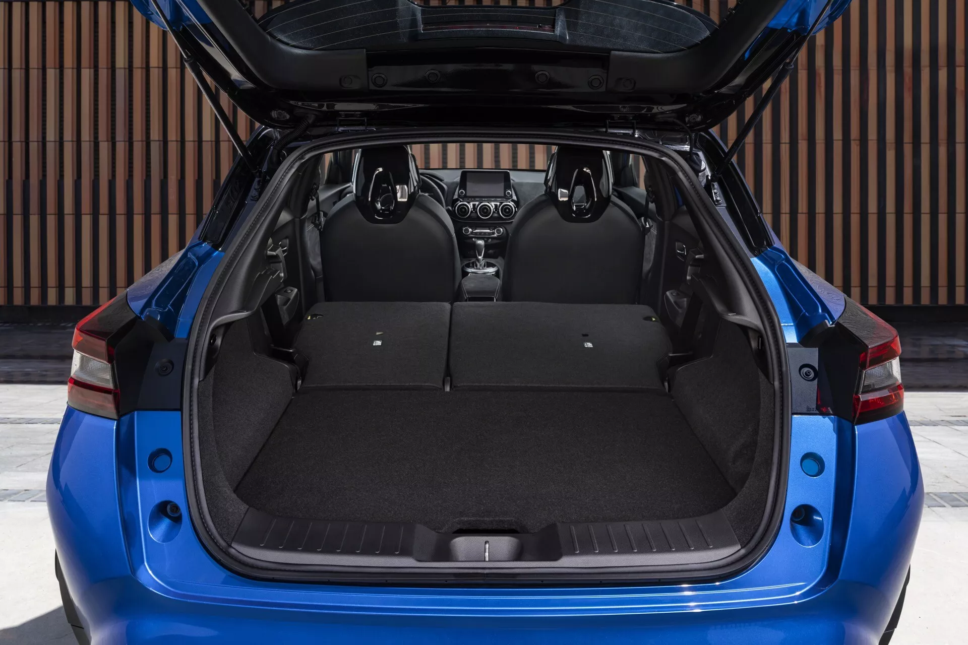 Nissan Juke F16 z roku 2019 v modré barvě, zadní pohled s otevřeným kufrem ukazující prostorný nákladní prostor, dělená zadní sedadla s možností sklopení a viditelný zatahovací kryt nákladu