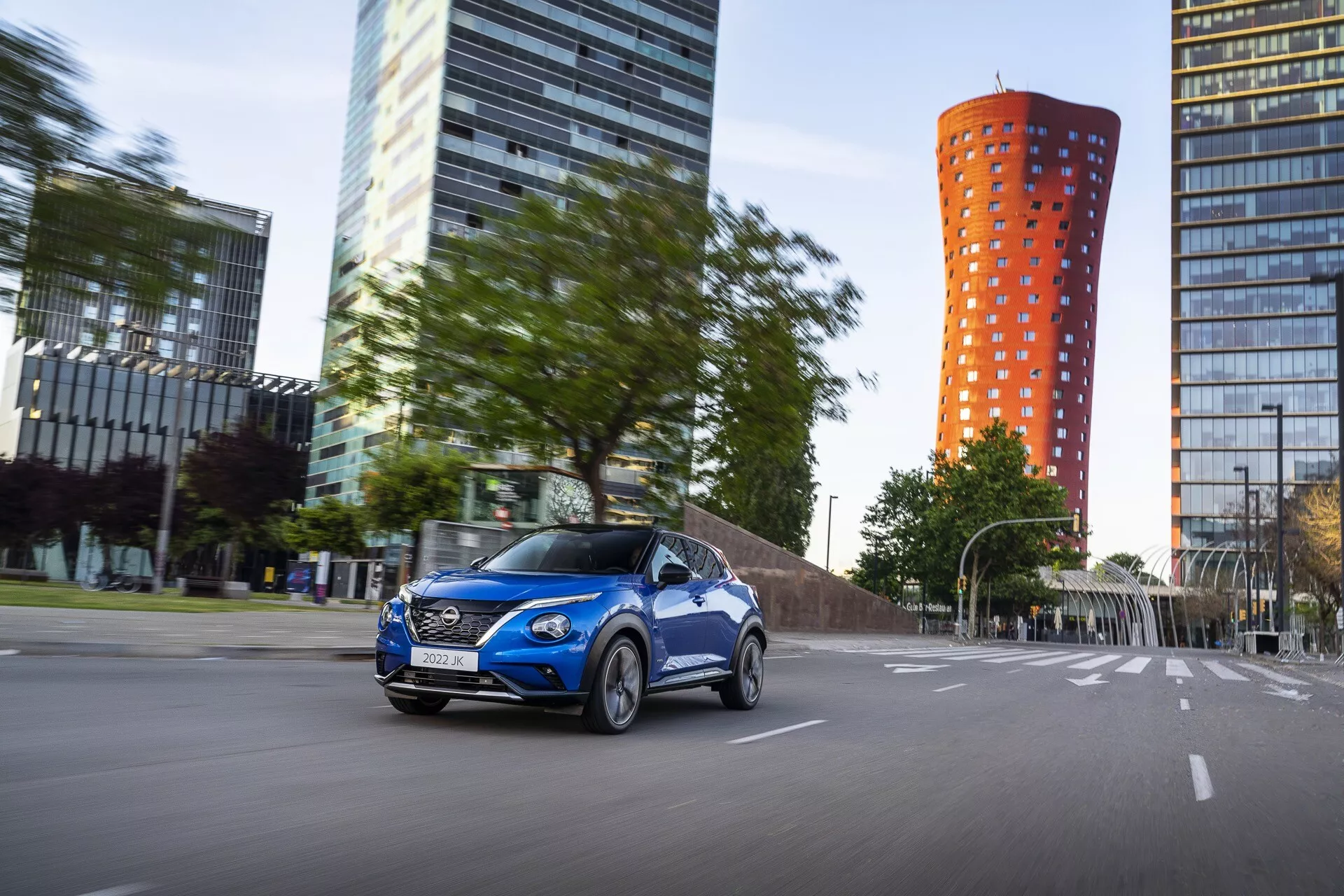 2019 Nissan Juke F16 ve výrazné modré barvě, v pohybu na městské silnici s dynamickým pohledem ze strany řidiče, představující slitinová kola, obrysové karosářské práce a zapnutá přední světla na pozadí moderního města s efektem rozmazání pohybu.