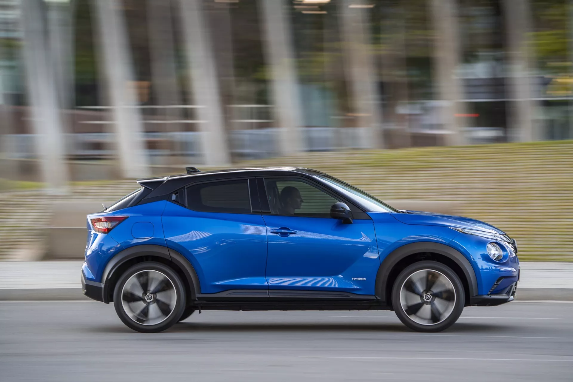 Modrý Nissan Juke (F16) 2019 s moderním designem a technologiemi jako jsou aerodynamické linie karoserie, slitinová kola a potenciální bezklíčový přístup, pohybující se dynamicky s rozmazáním pohybu v zamračený den
