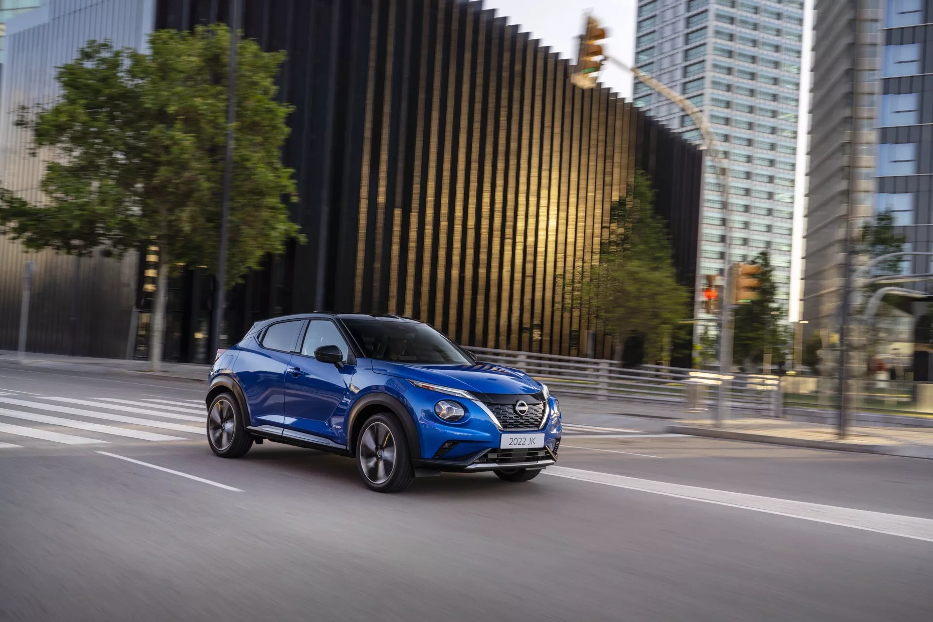 Moderní kompaktní SUV Nissan Juke (F16) z roku 2019 v pohybu, zachycené z boční perspektivy v jasně modré barvě, ukazující design vozidla, dynamickou přítomnost a aerodynamické prvky na pozadí města.