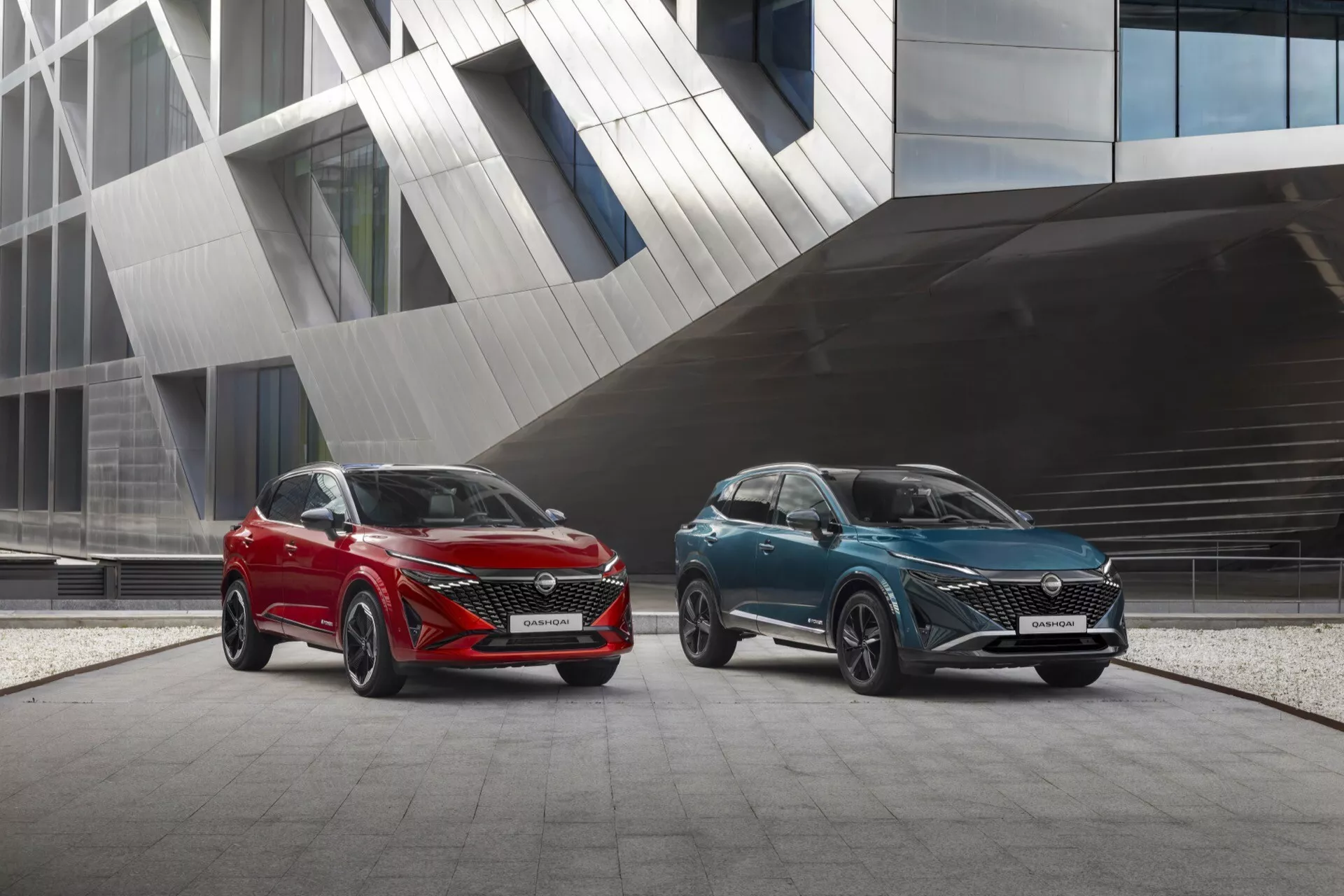 Obrázek dvou vozů Nissan Qashqai 2024 (J12-facelift) zaparkovaných vedle sebe před moderním architektonickým pozadím. Červené auto vlevo je v popředí a ukazuje svou přední pravou část s aerodynamickými liniemi, ostrými LED světlomety a výraznou maskou chladiče. Metalický modrý vůz vpravo zobrazuje svou přední levou část, která rovněž zdůrazňuje odvážné designové prvky a pokročilou osvětlovací technologii. Oba vozy působí prémiově a moderně, s možnými funkcemi jako autonomní řízení, pokročilé informační systémy a hybridní nebo elektrické pohony.