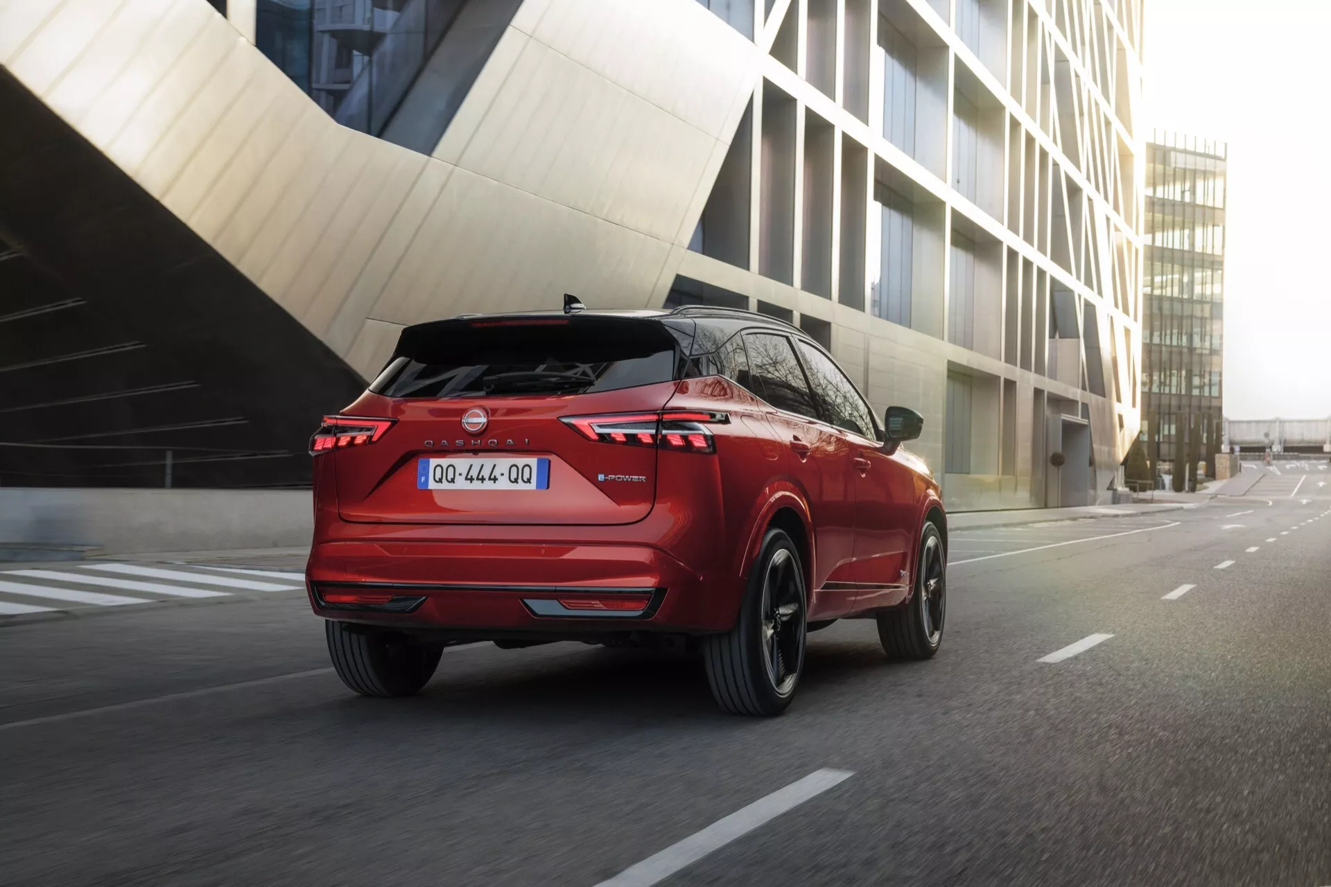 Nízký úhel, pohled na zadní tři čtvrtiny moderního SUV jedoucího po městské ulici, zdůrazňující jeho ostré linie, aerodynamický profil, rozsvícená zadní světla, dvoubarevné schéma s černým spodním nárazníkem, zadní spoiler, středovou SPZ a litá kola.