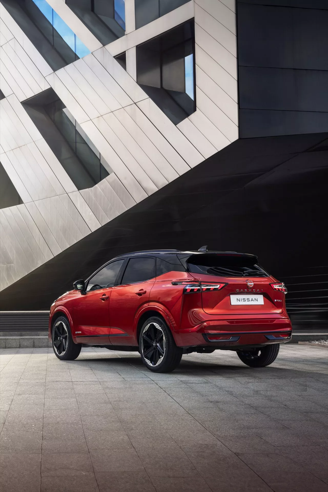Zadní tři čtvrtinový pohled na červený SUV Nissan Qashqai 2024 (J12-Facelift), který předvádí jeho levou stranu, zadní část a část střechy v městském prostředí s moderní architekturou. Obrázek zvýrazňuje vodorovná zadní světla vozidla, černá vícepaprsková kola z lehké slitiny, vzor běhounu pneumatik, čistě navržený zadní nárazník s integrovanými parkovacími senzory nebo odrazkami a lesklý lak. Na automobilu je jasně vidět značka s logem Nissan a názvem modelu.