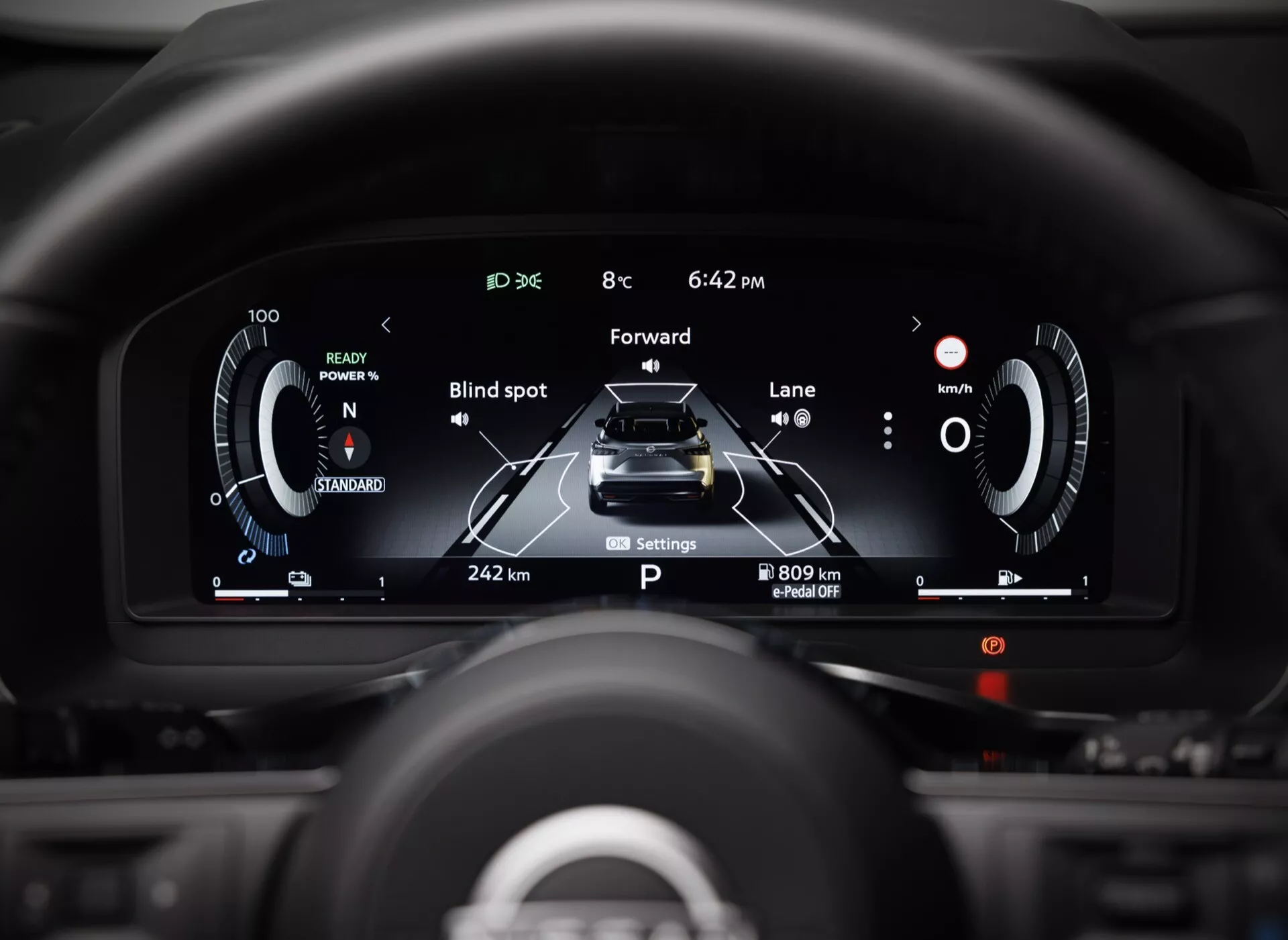 Detailní pohled na digitální přístrojovou desku vozu Nissan Qashqai 2024 z pohledu řidiče, zobrazující vysoké rozlišení displeje s prvky jako tachometr, otáčkoměr, ukazatel spotřeby/napájení, animovanou grafiku vozu s ukazateli asistenčních systémů řidiče jako sledování mrtvého úhlu a asistence udržování v jízdním pruhu, palubní počítač zobrazující vzdálenost a odhadovaný dojezd, stav vozidla včetně režimu 'Park' a upozornění na otevřené dveře, a aktuální čas a venkovní teplotu.