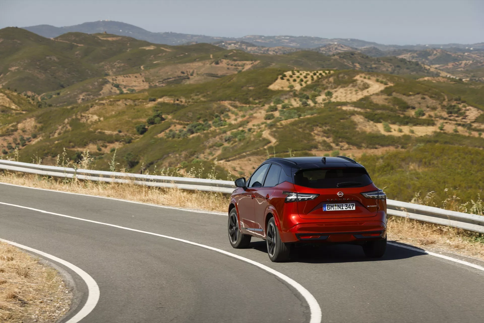 Živý červený Nissan Qashqai 2024 (J12-facelift) zachycený z tříčtvrtečního zadního pohledu při jízdě po malebné horské silnici. Zvýrazněny jsou zadní a pasažérské strany, jako jsou zadní světla, zadní nárazník, výfuk, zadní okno a značení modelu a značky, což ukazuje design vozidla a dynamický pohyb za jasného denního světla.