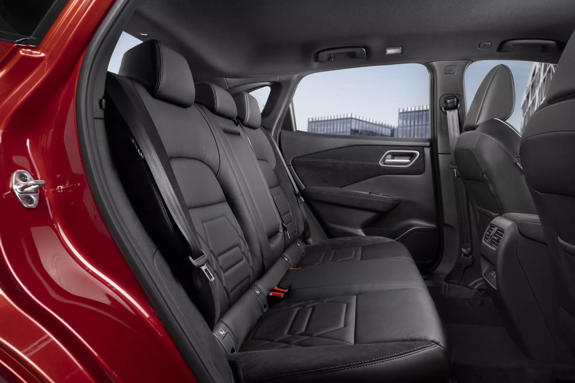 Interiér zadního pasažérského prostoru ve voze Nissan Qashqai 2024 J12-Facelift, zobrazující černé kožené sedadla lavicového stylu se středovou sklopnou loketní opěrkou a držáky nápojů, viditelné bezpečnostní pásy a opěrky hlavy pro tři pozice sezení, otevřené dveře vozu odhalující ovládání elektrických oken, reproduktor a úložný prostor, chromované akcenty na klice dveří a částečný pohled na klimatizační systém pro zadní pasažéry na zadní straně středové konzole.