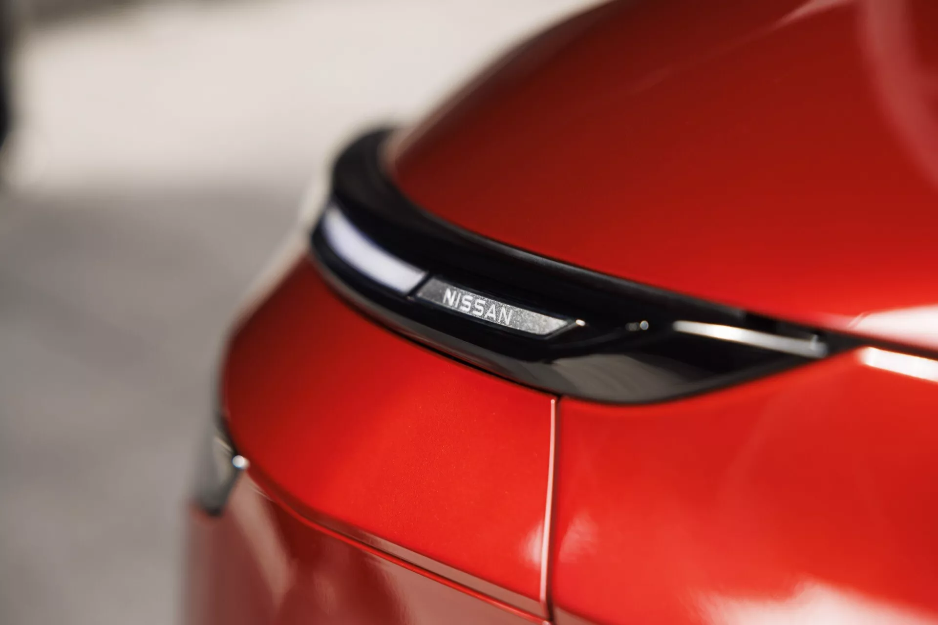 Detailní vysoké rozlišení fotografie zadní části červeného Nissan Qashqai 2024 (J12 Facelift), zaměřující se na centrální emblém nad zadní světelnou sestavou, s lesklým černým ozdobným prvkem a dynamickým záhybem na kufru, s reflexní lesklou barvou a hladkým bokeh efektem.