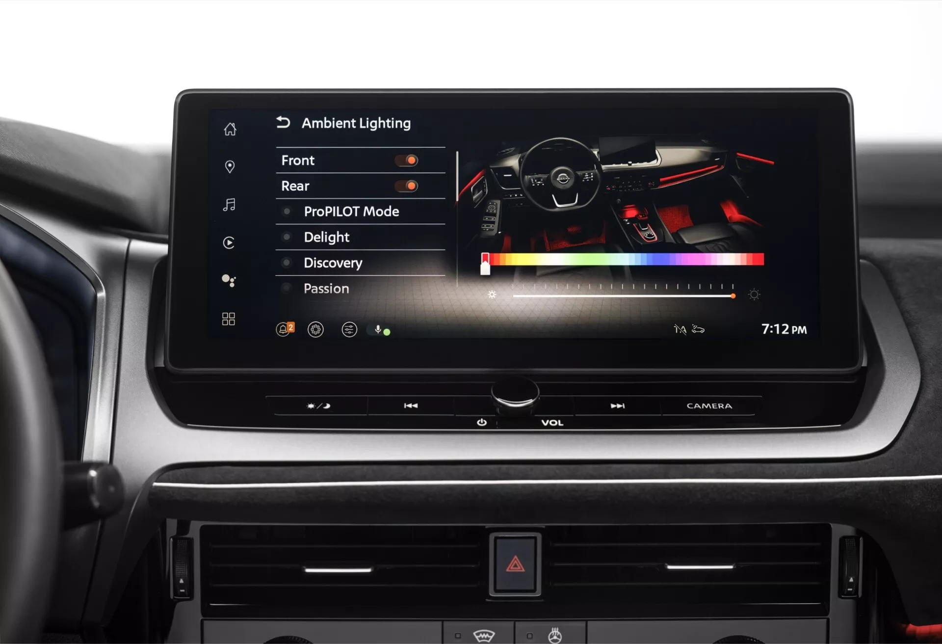 Pohled na interiér Nissanu Qashqai 2024 J12 Facelift zaměřený na centrální palubní desku. Pohled zepředu zdůrazňuje informační systém s aktivním širokým, horizontálně orientovaným dotykovým displejem zobrazujícím nabídku 'Ambientní osvětlení'. Displej je ostrý s černým pozadím a bílým textem, ukazuje možnosti výběru barev a odráží interiér vozu, včetně volantu. Níže jsou fyzické ovladače se dvěma knoflíky pravděpodobně pro hlasitost a ladění, řada tlačítek pro audio a klimatizaci, a centrálně umístěné tlačítko výstražných světel. Informační systém nabízí přehrávání zvuku, nastavení vozidla a připojení smartphonu, moderní design vozu s nastavitelným ambientním osvětlením a hmatatelným digitálním rozhraním.