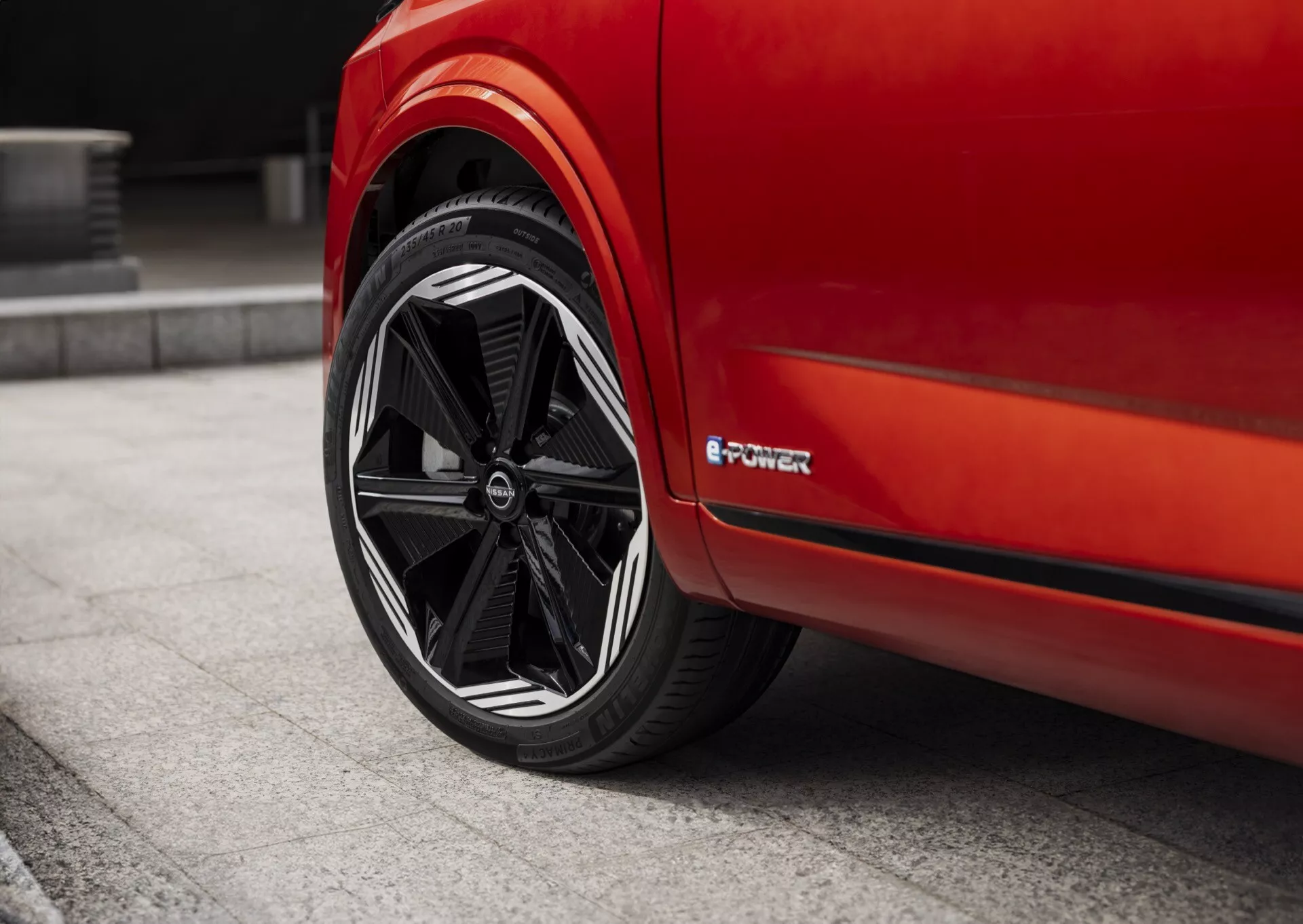 Detailní pohled na zadní kolo a boční část karoserie červeného Nissan Qashqai 2024 (J12-facelift) z úrovně země. Kolo má vícepaprskový design s černým povrchem a kontrastním detailem vnějšího ráfku. Složitý hvězdicový vzor paprsků vychází z centrálního náboje, který nese logo. Ráfek má odznak 'e-POWER', který značí elektrickou nebo hybridní technologii vozu. Nízkoprofilová pneumatika zdůrazňuje sportovní výkon. Boční část karoserie je hladká bez viditelných klik dveří nebo ozdob, což zdůrazňuje aerodynamický vzhled.