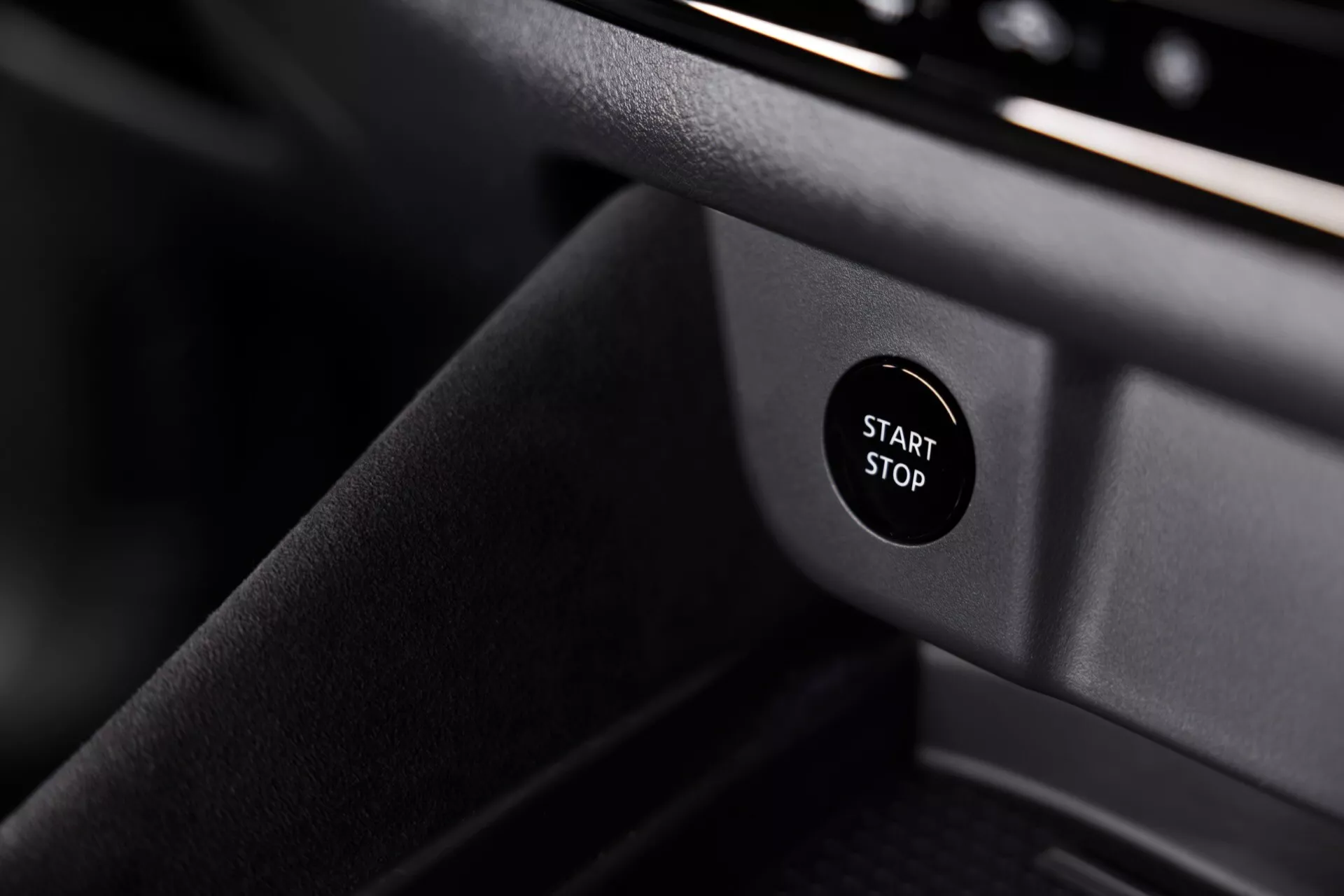 Pohled zblízka na interiér Nissan Qashqai 2024 (J12-facelift), který zdůrazňuje tlačítko start-stop na palubní desce. Tlačítko s bílým textem 'START STOP' na černém pozadí je ostře zaostřeno proti tmavému, měkce rozmazanému pozadí, což je typické pro moderní, elegantní design vozidla.