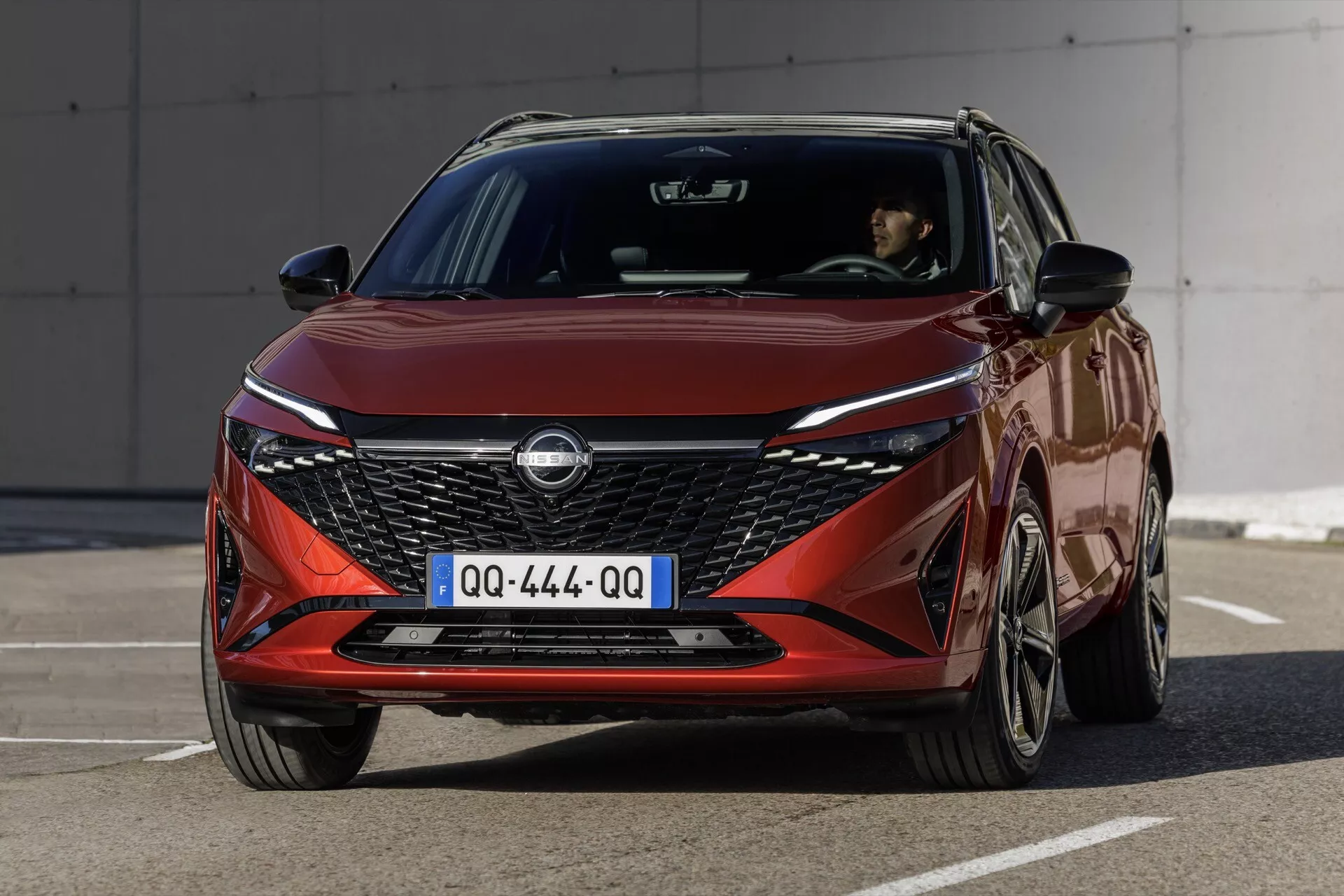 Fotografie vozu Nissan Qashqai 2024 (J12-Facelift), pořízená z předního tříčtvrtečního pohledu. Vozidlo v metalické barvě hluboké červené interaguje s osvětlováním, zdůrazňující V-tvarovanou mřížku chladiče, elegantní světlomety, tvarované linie kapoty vedoucí k centrálně umístěnému emblému a víceramenné slitkové ráfky kol. Vozidlo je umístěno na dlážděném povrchu bez překážek, což zvýrazňuje moderní a agresivní design vozu se zaměřením na funkce, jako jsou denní LED světla umístěná ve světlometech.
