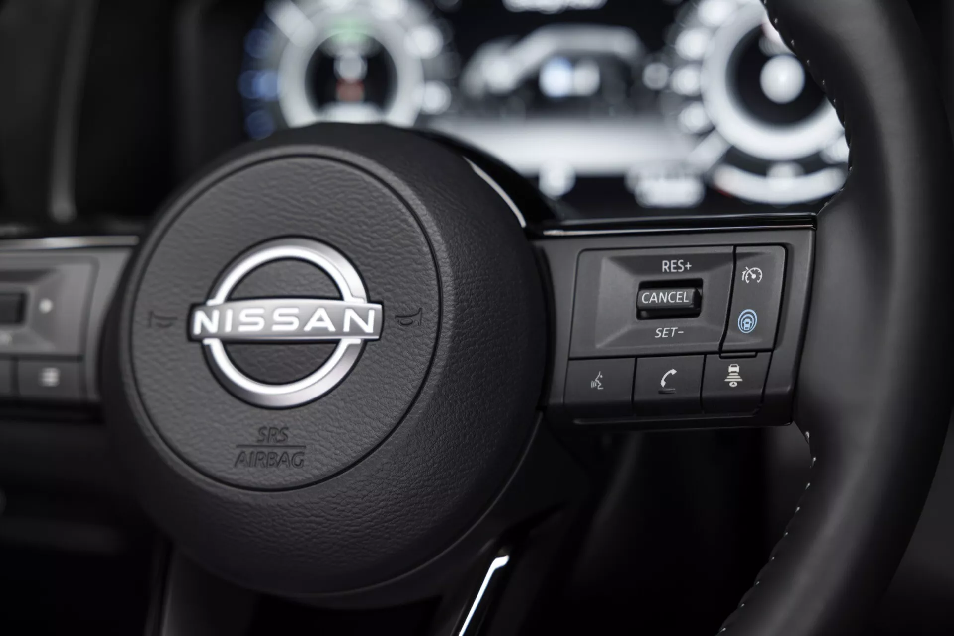 Detailní pohled na volant modelu Nissan Qashqai 2024 (J12-facelift), zaměřený na ovládací prvky na levé straně a logo značky. Vysoce detailní obrázek zvýrazňuje tlačítka pro tempomat, hands-free volání a bezpečnost při kolizi, usazené na volantu obaleném černou kůží. Rozostřené pozadí zahrnuje část přístrojové desky se svítícími ukazateli, zdůrazňující ergonomický design a pokročilé technologické funkce vozu.