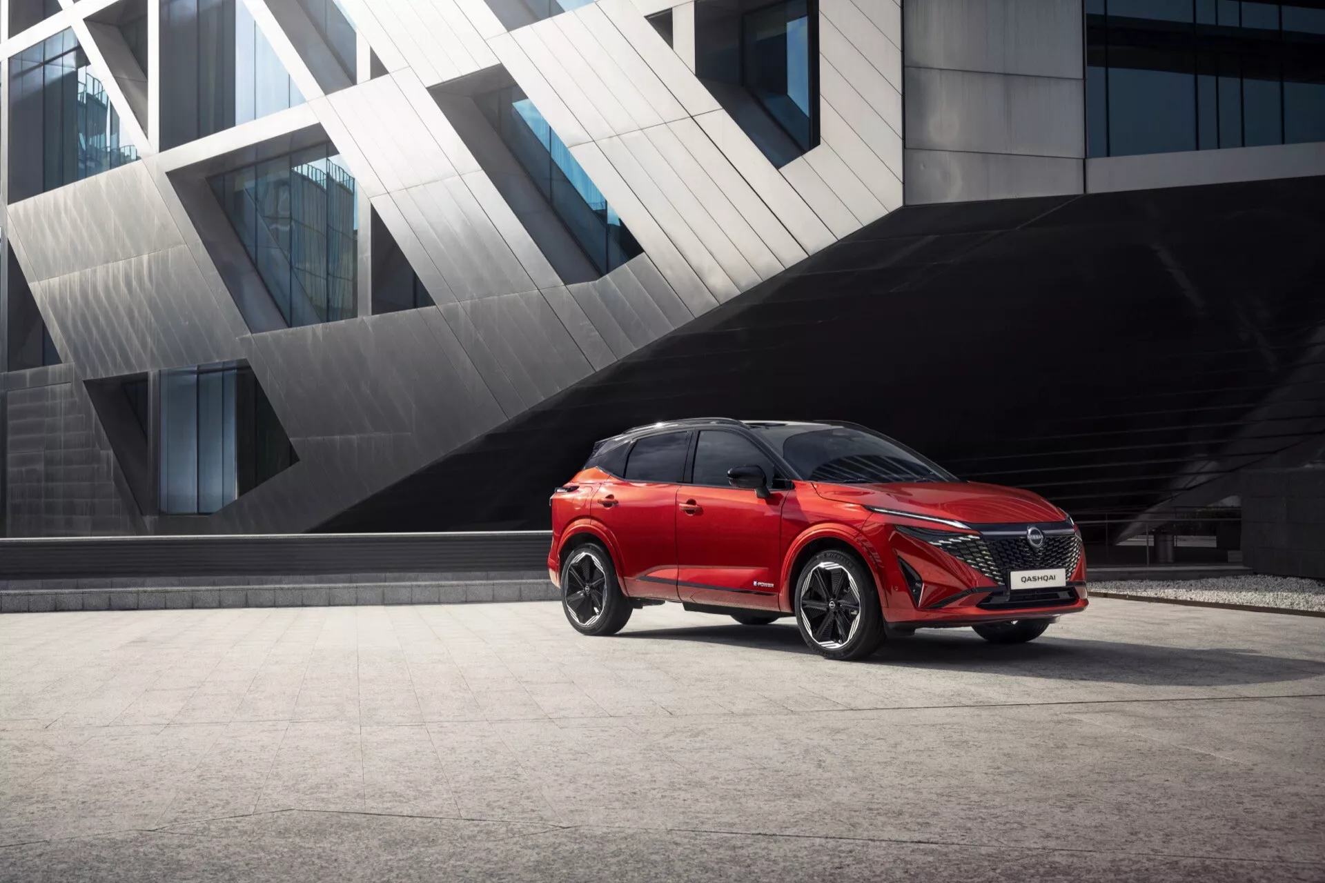 2024 Nissan Qashqai (J12-Facelift) v zářivě červené barvě je vyobrazen zaparkovaný proti městskému architektonickému pozadí. Geometrické struktury, možná moderní budova, jsou součástí scény. Obrázek zachycuje nízkoúhlý, tříčtvrtinový přední pohled na auto a ukazuje přední mřížku, LED světlomety, dveře na straně řidiče, boční zrcátko, přední kolo a část střechy. Klíčové prvky jako pokročilé LED světlomety, aerodynamické linie karoserie, velká hliníková kola, boční zrcátko s integrovanou technologií a výrazná přední mřížka naznačují technologickou sofistikovanost a moderní design auta, které vyzařují prémiový dojem.