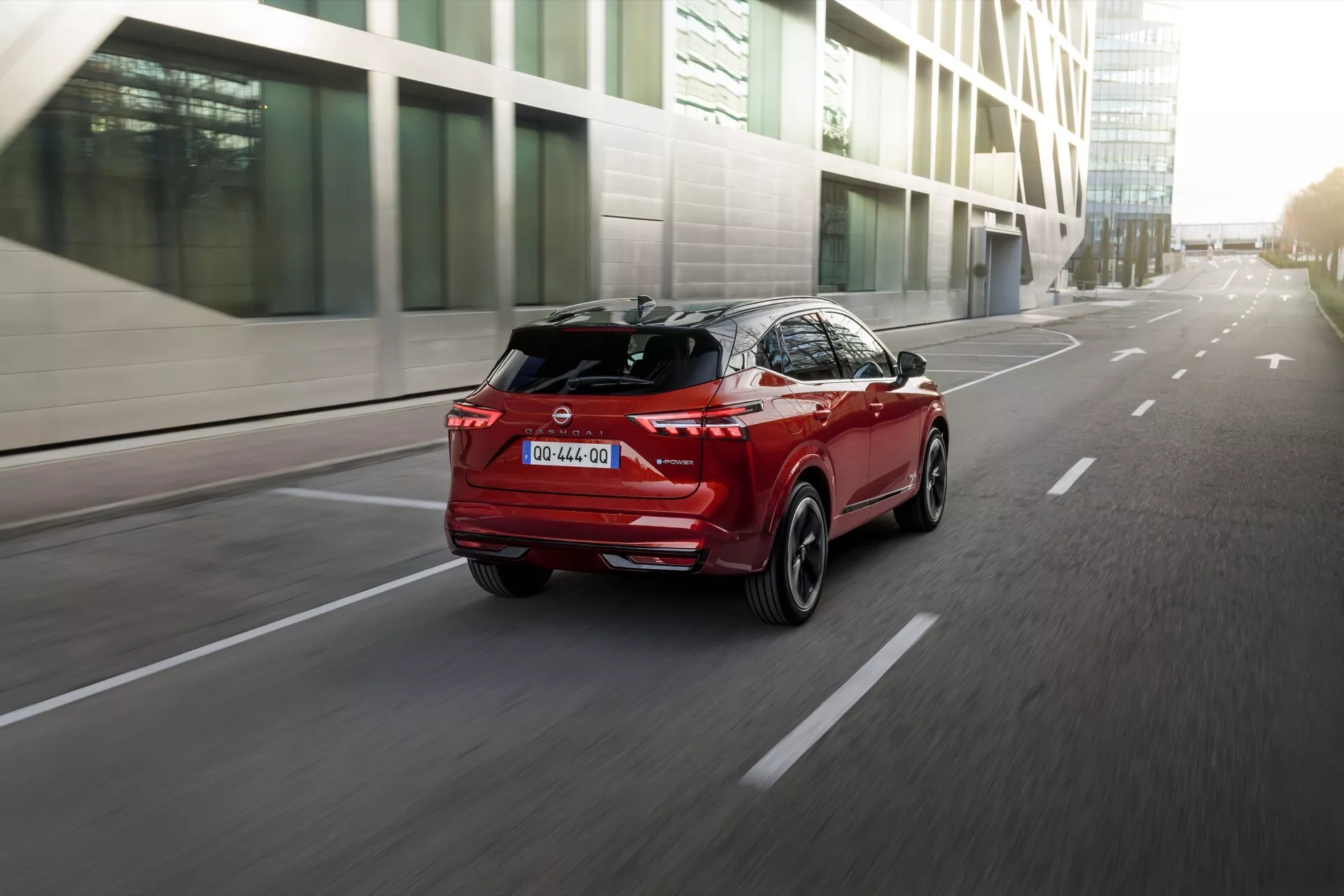Dynamický záběr zadních tří čtvrtin červeného SUV Nissan Qashqai 2024 J12-Facelift jedoucího po asfaltové silnici, se zřetelně osvětlenými ostrými úhlovými zadními světly, výrazným zadním spoilerem a zadním difuzorem. Mírně nízký úhel záběru zvýrazňuje výšku a postoj vozu, se zřetelnou žraločí ploutví na střeše a chromovaným lemem podél spodních částí dveří. Moderní městské prostředí v pozadí je rozmazané, čímž je zdůrazněn sportovní charakter a pokročilý design SUV.
